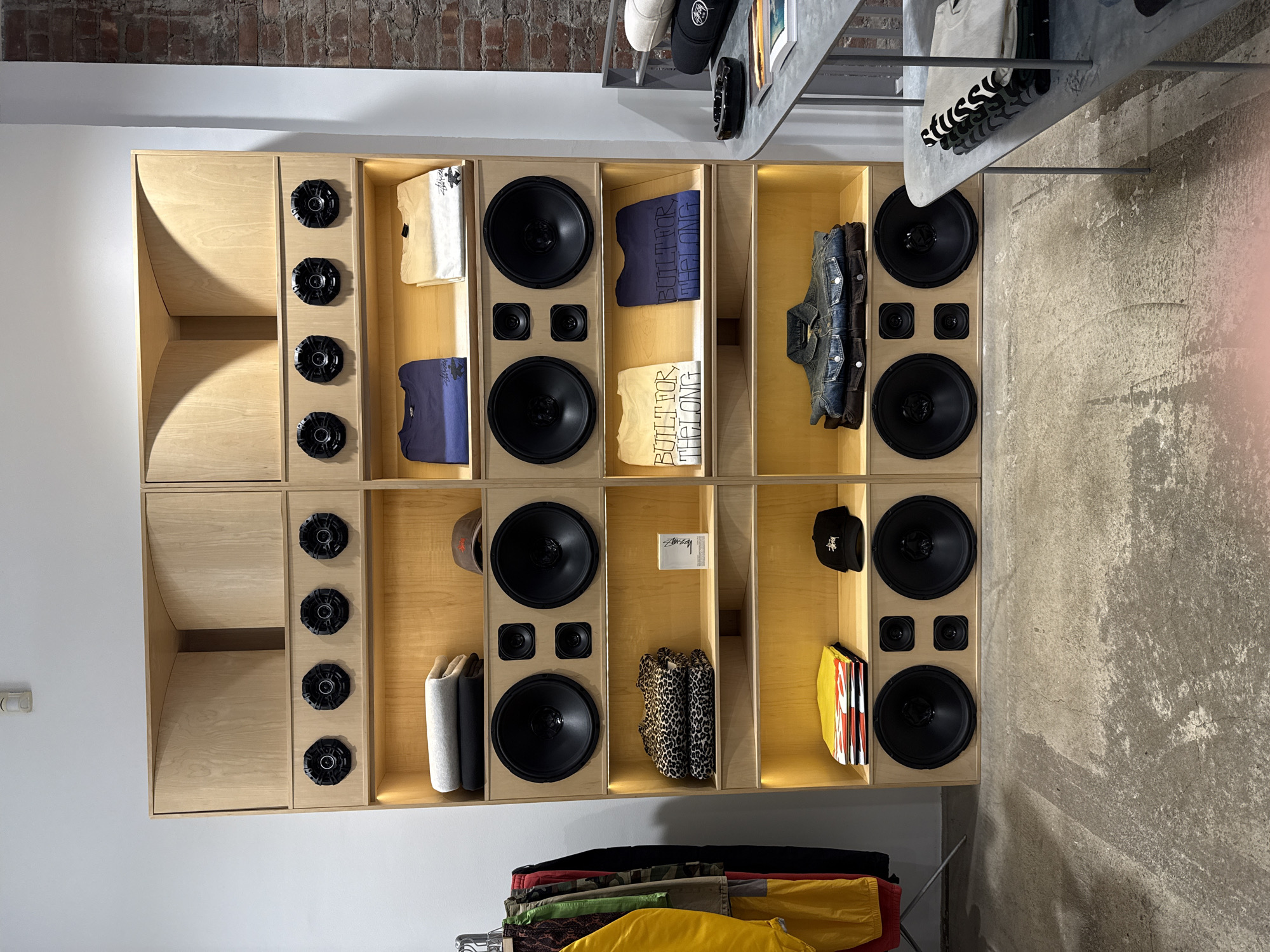 The image depicts the interior of a retail store located in the city of New York, United States of America. A large, custom-built display unit, fabricated from light-colored wood, occupies the central and left portions of the frame. This unit is vertically segmented into two columns and horizontally into multiple levels, designed to resemble a large-scale sound system.
The uppermost sections of the wooden unit feature curved, horn-shaped enclosures. Below these, eight small, black circular speakers are recessed into the wood (four per column). The next level down contains illuminated shelves displaying folded garments: grey and black sweaters/sweatpants on the left, and navy blue and light tan Stussy-branded t-shirts on the right, accompanied by a brown baseball cap.
The central portion of the unit integrates four large, black circular woofers and eight smaller circular speakers (two small speakers above each large woofer). Beneath these speakers, additional illuminated shelves are present. The left shelves hold stacked leopard-print garments and a white Stussy-branded product box, with stacked multi-colored garments and a black Stussy baseball cap below. The right shelves display folded light tan and navy blue t-shirts featuring the text "BUILT FOR THE LONG," with a stacked denim jacket over a brown jacket below. The bottom section of the wooden unit mirrors the central speaker configuration with four large woofers and eight smaller speakers.
To the immediate left of the wooden unit, a metal clothing rack holds various garments, including yellow and red outerwear, camouflage-patterned pants, and dark trousers.
On the right side of the image, an exposed red brick wall is visible behind a multi-tiered metal display table with rounded edges. The top tier of this table holds a black ceramic ashtray, a white rolled item, and a black hat. The lower tiers display stacked folded garments, including items with a black-and-white striped pattern, and several books or magazines.
The floor is a distressed concrete surface with varying shades. The walls are predominantly white, with a white smoke detector mounted high on the left wall. No people are present in the scene, and no actions or interactions are depicted.