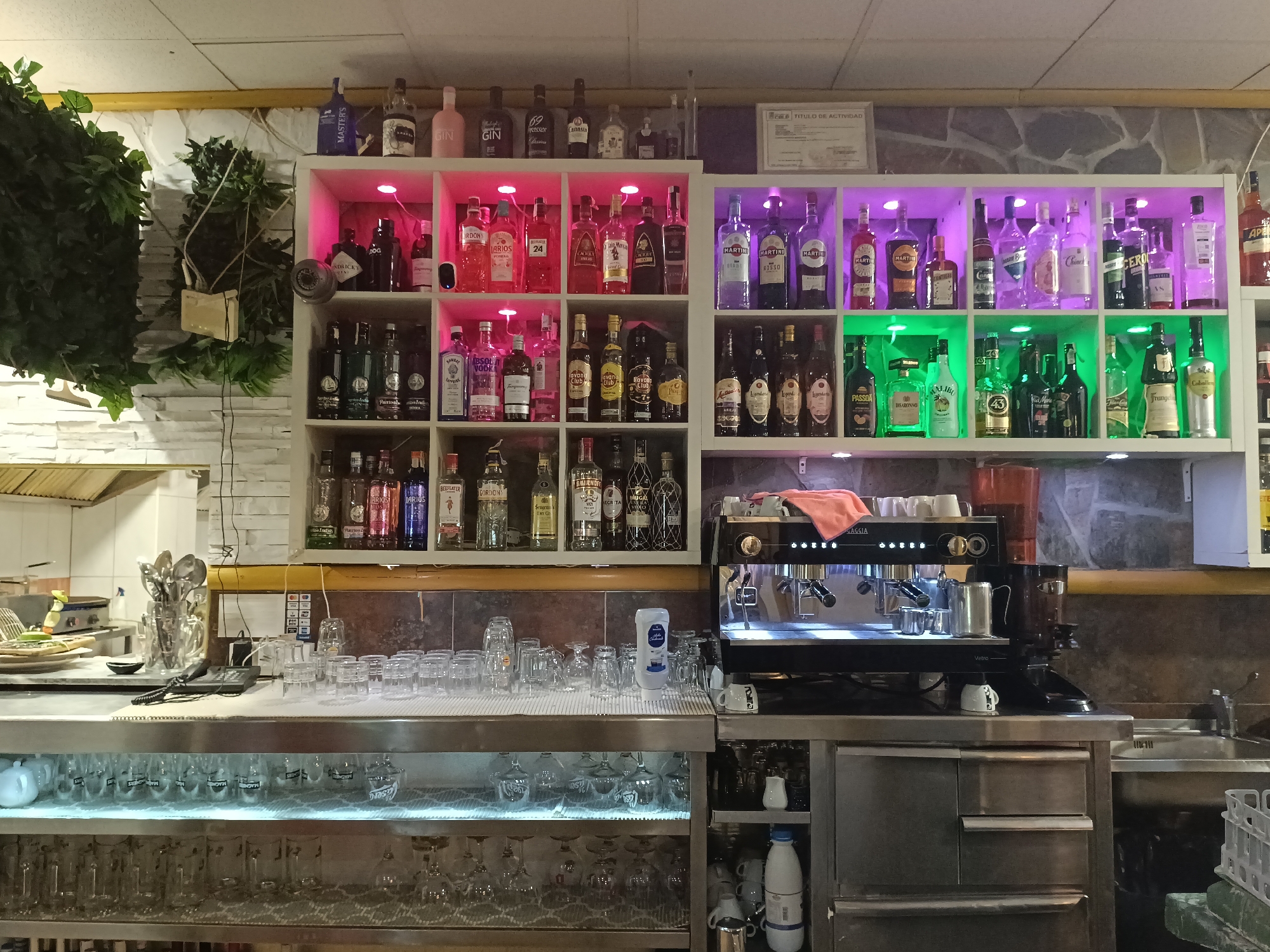 The image shows the interior of a bar or restaurant in San Gabriel, Spain. The scene is dominated by shelves stacked with various bottles of liquor and other beverages. On the top shelf, several bottles of different brands are visible, including gin and vodka. Below this, illuminated by pink and purple lights, are more bottles of liquor, some appearing to be cocktails or liqueurs. The middle shelves also feature rows of bottles, including brands like "Havana Club" and various types of spirits. The bottom shelves display an assortment of glassware, some of which appear to be wine glasses and tumblers.
In the foreground, a stainless steel counter holds a collection of clean glasses, a coffee machine, and a small pile of cleaning supplies. Behind the counter, kitchen equipment, including what appears to be a grill or stove, is partially visible. The overall environment suggests a well-stocked establishment ready for service. There are no people visible in the image. The lighting in the bar is varied, with spotlights illuminating the shelves of bottles. The background shows a stone wall and what looks like a certificate or diploma hanging on the wall.
