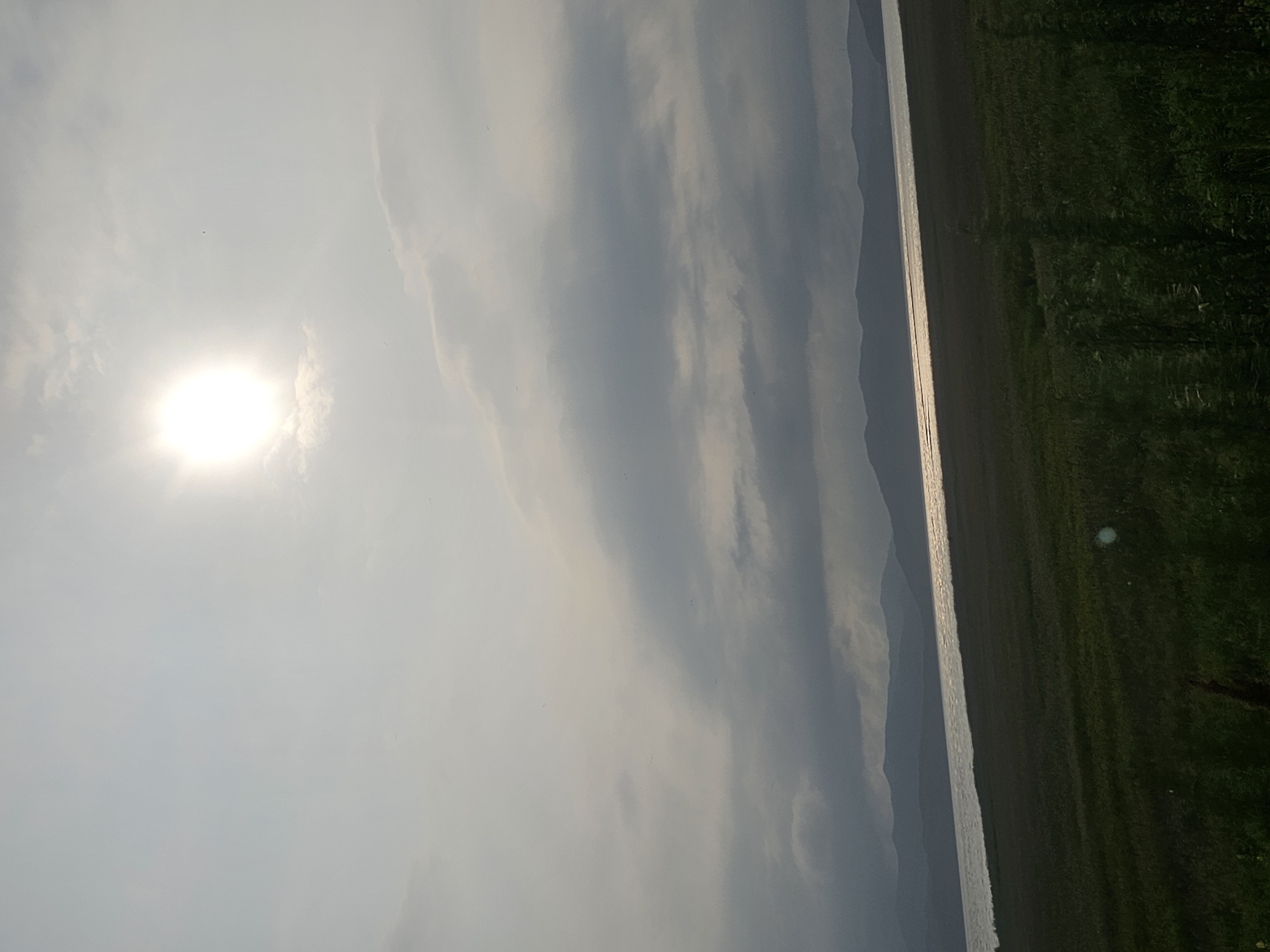 The image depicts a landscape scene at a body of water with distant hills under a cloudy sky. The sun is visible as a bright, diffused orb in the upper portion of the frame, casting a shimmering reflection on the water. The foreground consists of a grassy field. The scene is captured in a low-light, possibly overcast, condition. The location is identified as Shkotovo, Russia.