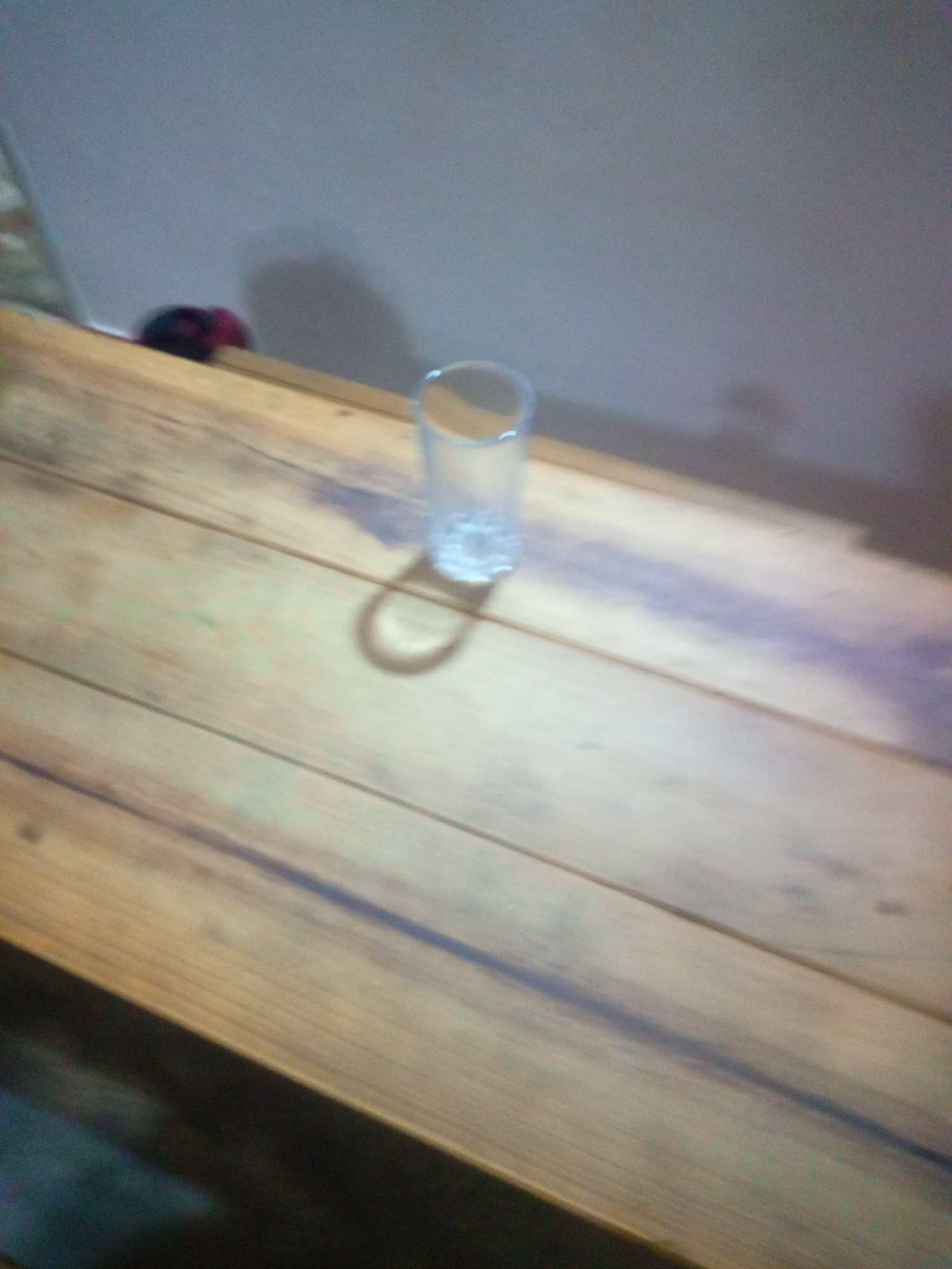 In Altagracia, Venezuela, a clear glass is positioned on a wooden surface, casting a shadow. The wooden surface appears to be a table or countertop, with visible grain and plank lines. The background is a wall, partially illuminated, with a darker area suggesting a shadow or obscured region. To the left, a blurred object with dark and reddish hues is partially visible, possibly clothing or fabric. The overall lighting is dim, contributing to the indistinct nature of some details.