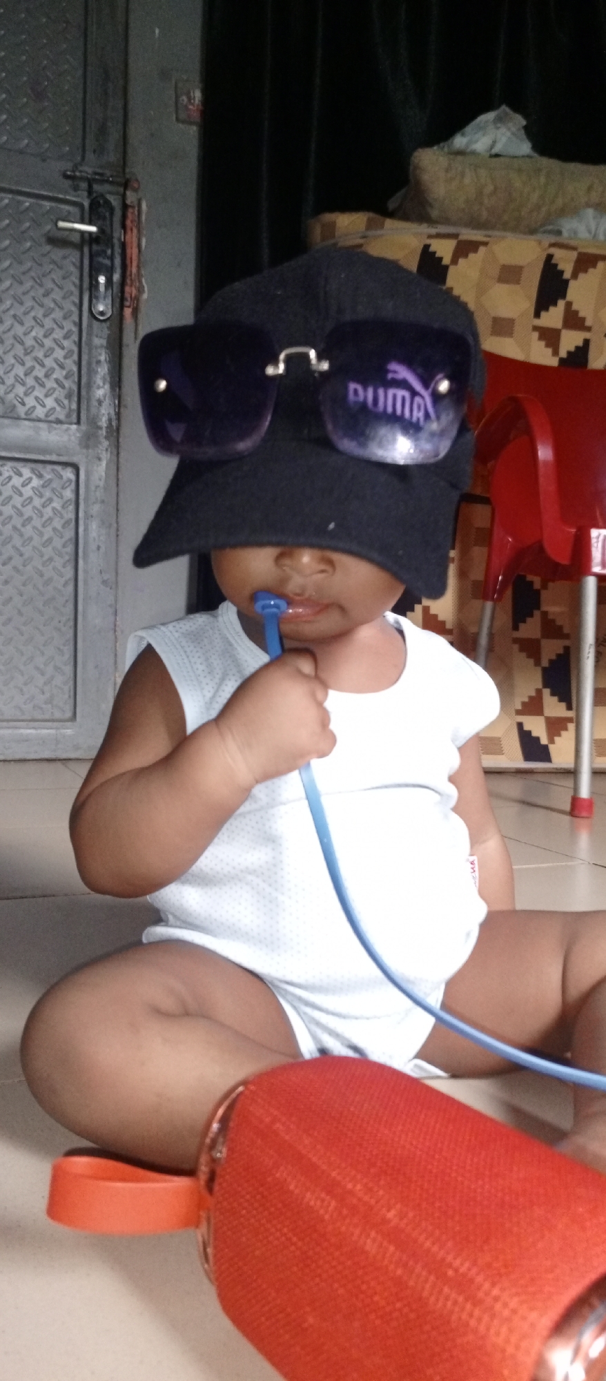 A baby is sitting on the floor wearing a white shirt, a black hat, and sunglasses with the word "PUMA" on them. The baby is holding a blue object in their mouth. In front of the baby is a red cylindrical object. Behind the baby are a door, curtains, a chair, and a patterned surface. The location is Unknown, Unknown.