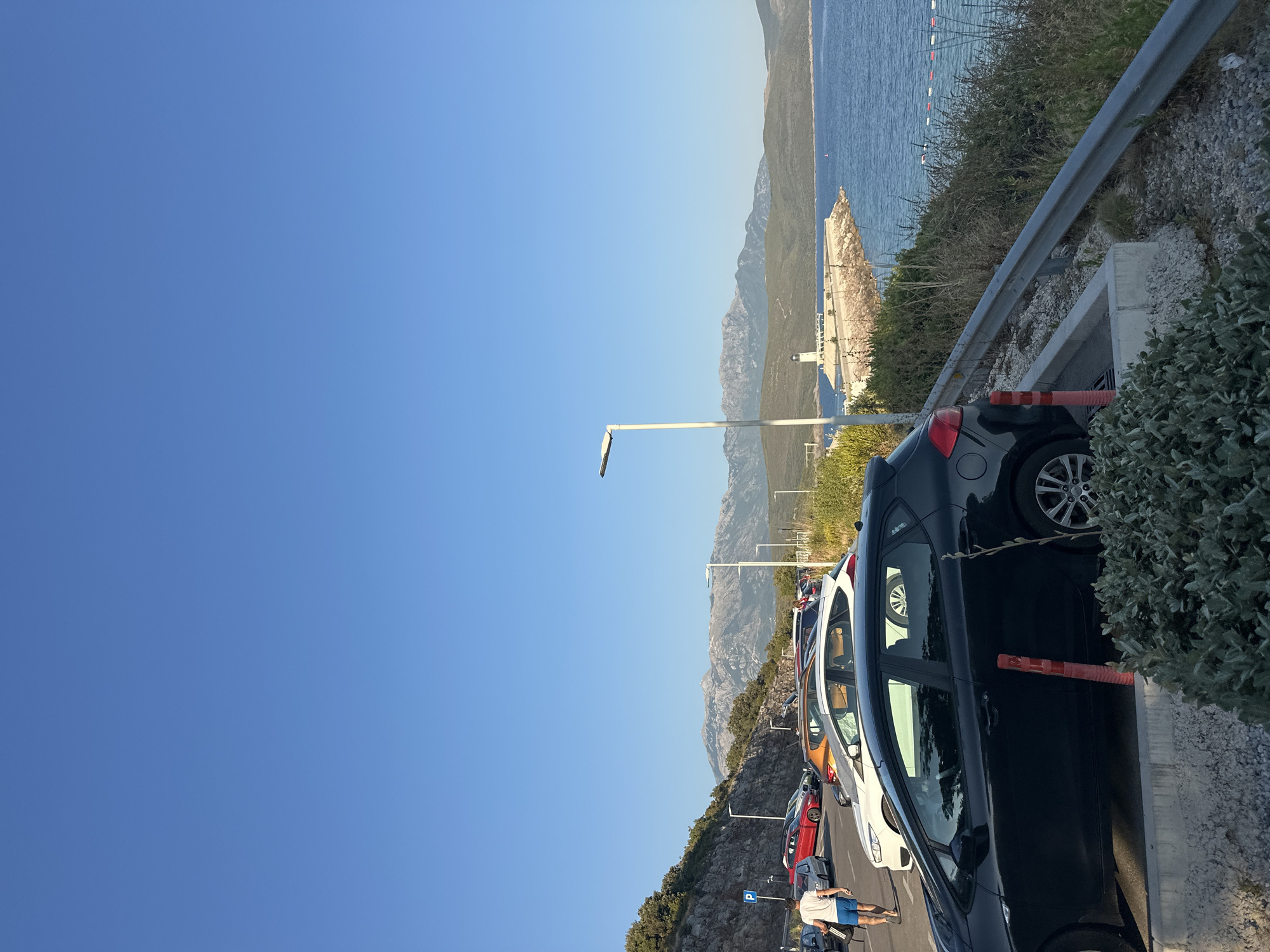 The image depicts a coastal parking lot near Radovići, Montenegro, on a clear, sunny day with a bright blue sky. In the foreground, a black hatchback car is parked, partially obscuring a red traffic cone. Adjacent to the car is a concrete parking barrier and gravel. Beyond the car, a line of other vehicles, including a white hatchback and a red sedan, are parked on a paved road that curves along the base of a rocky hillside covered in sparse vegetation. A person in blue shorts and a white t-shirt is walking away from the viewer towards the parked cars. Further in the background, a calm, blue sea stretches out, meeting a distant shoreline with rolling green hills and rugged mountains under the horizon. A white lighthouse stands prominently on a stone jetty extending into the sea. The scene is peaceful, with no apparent activity beyond the parked cars and the lone pedestrian.