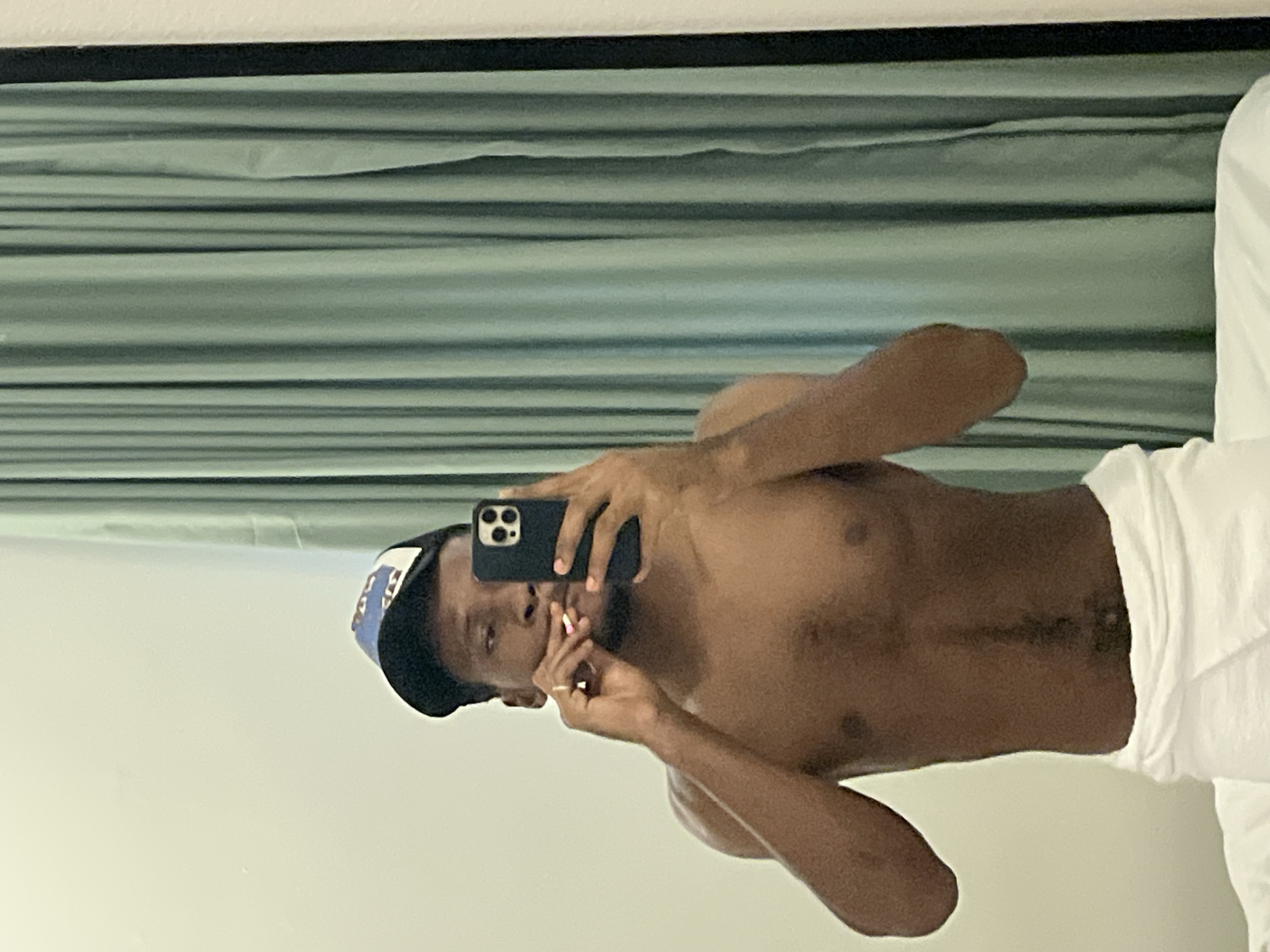 A dark-skinned male individual is depicted taking a mirror selfie indoors. He is shirtless, displaying his bare chest and torso, and has a white towel or garment wrapped around his waist. He wears a dark baseball cap with blue and white accents; the text or logo on the cap is not clearly legible.
The subject is holding a dark-colored smartphone, possibly an iPhone, with his left hand, pointing it towards the mirror to capture his reflection. His right hand is raised towards his mouth, holding a slender, light-colored object that appears to be a lit cigarette or joint. He is looking directly into the phone's camera with a neutral expression.
The background shows light blueish-gray or green horizontal blinds or curtains, suggesting a window covering. The surrounding walls are plain and light-colored, indicating an indoor setting. The scene conveys a casual moment, likely in a private room or residence in Gwarinpa, Nigeria. There is no clearly visible text apart from indistinct patterns on the cap.