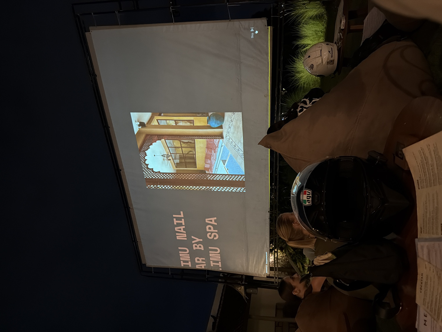 The image depicts an evening scene in an outdoor or semi-outdoor setting, likely a casual lounge or bar in North Kuta, Indonesia. A large white projection screen dominates the left side, illuminated against the dark sky.
Several people are present, though mostly obscured. One person with blonde hair is visible behind a black "AGV" motorcycle helmet, and another person with darker hair is partially seen in the lower right, both seemingly seated on brown bean bag chairs. Another white "SHARK" motorcycle helmet rests on a bean bag in the upper right. Paper menus, faintly showing "DRINKS," "COCKTAIL," and "MENU," are on a table to the right. Green foliage is visible in the background, adding to the outdoor ambiance.
The main activity is the viewing of a video projection on the large screen. The screen displays an image of an interior with traditional arched windows and a water feature, possibly from a spa or resort. Text projected vertically on the screen reads "IMU NAIL BAR BY IMU SPA." In the top left corner of the projection, a "SKIP" button with a play icon is visible. The overall scene suggests a relaxed and casual outdoor movie night or presentation.