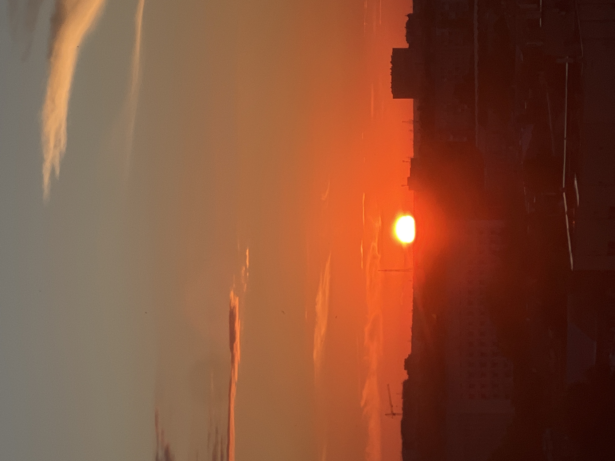 This image captures a vibrant sunset over the city of Odesa, Ukraine. The sky is a rich tapestry of oranges and reds, with streaks of pink and yellow from scattered clouds. The sun, a brilliant, almost blinding orb, is setting on the horizon, casting a warm glow over the cityscape. The silhouette of buildings and what appears to be a construction crane are visible against the dramatic sky, hinting at the urban environment of Odesa. The overall impression is one of natural beauty and urban development coexisting.