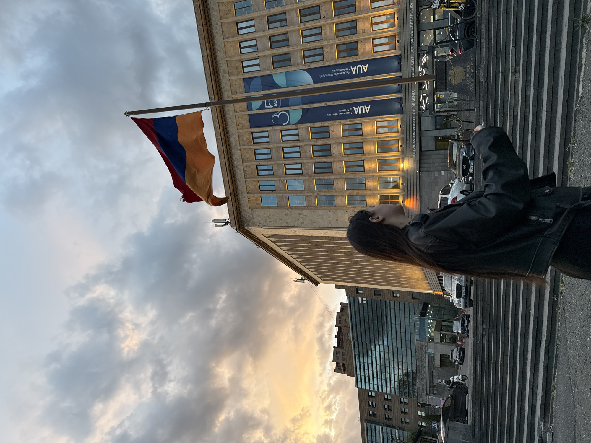 The image shows a street scene in Yerevan, Armenia, featuring the American University of Armenia (AUA). The Armenian flag is prominently displayed in the foreground, waving against a cloudy sky. The AUA building is visible behind the flag, adorned with banners. A person is visible on the right side of the frame, looking upwards toward the flag. A modern glass building can also be seen in the background. The foreground includes stairs and parked cars, indicating a public urban area. The overall ambiance suggests a vibrant city environment, with a combination of modern architecture and national symbols.