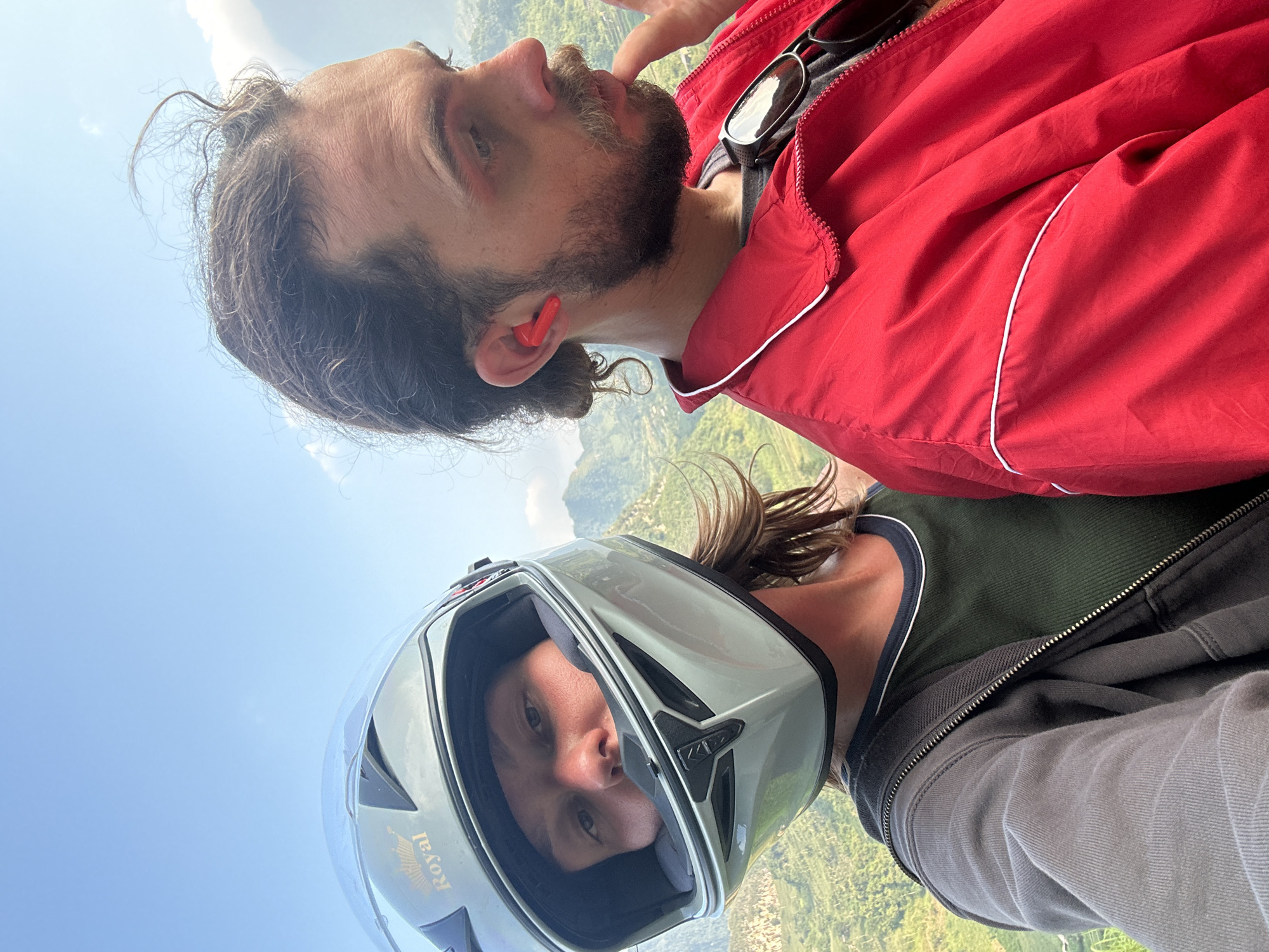 Two people are posing for a selfie. The first person is wearing a red jacket, has a beard, and an orange earbud in their left ear. Sunglasses are placed on the jacket. The second person is wearing a helmet and a green shirt and a gray jacket. The background suggests a mountainous or hilly terrain, possibly taken during paragliding. The image appears to be taken in Du Già Commune, Vietnam. The sky is visible in the background.