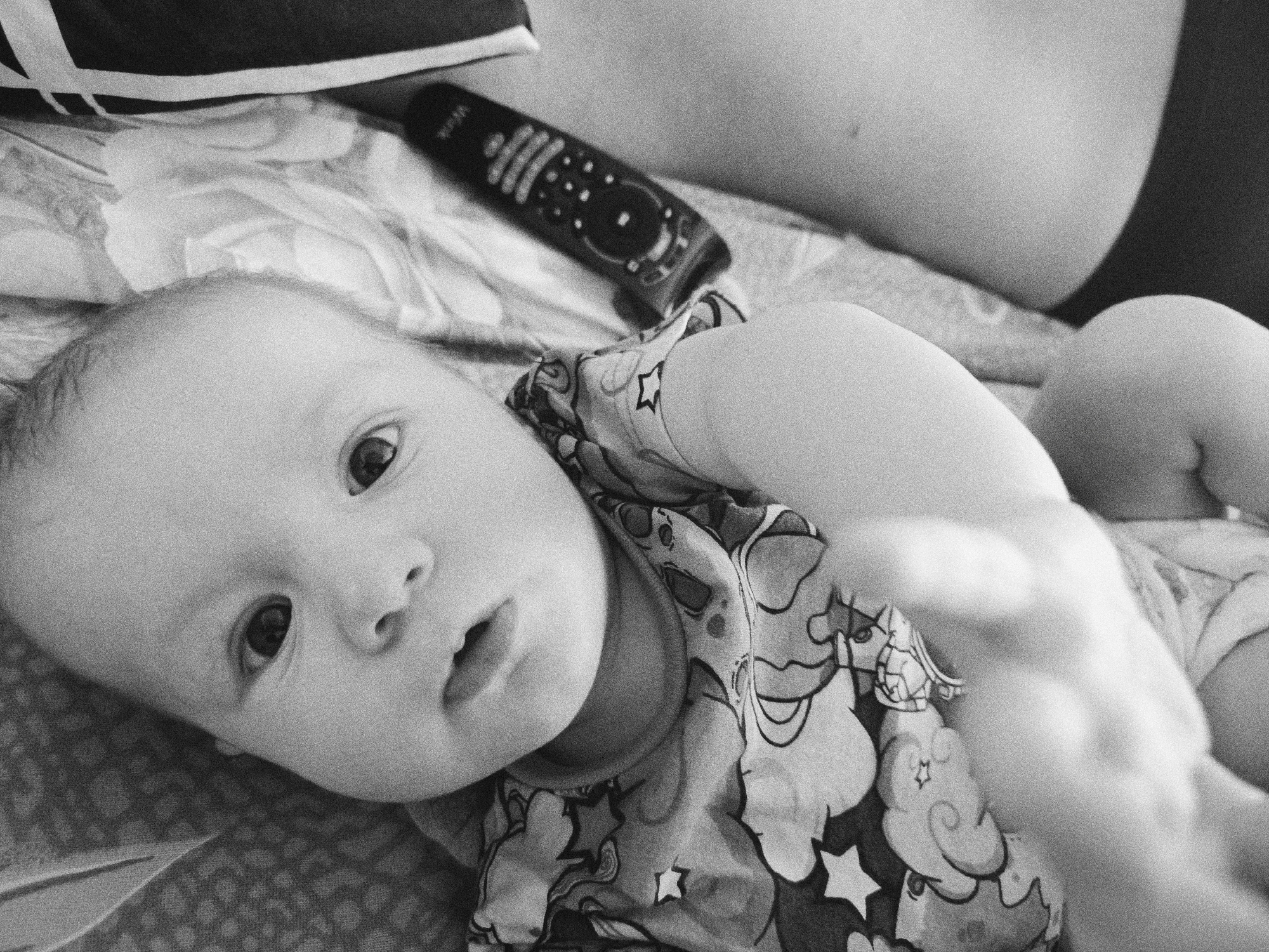 A black and white photograph depicts a baby lying on a patterned fabric. A remote control is near the baby's head. A person lies to the right. The location is unknown.