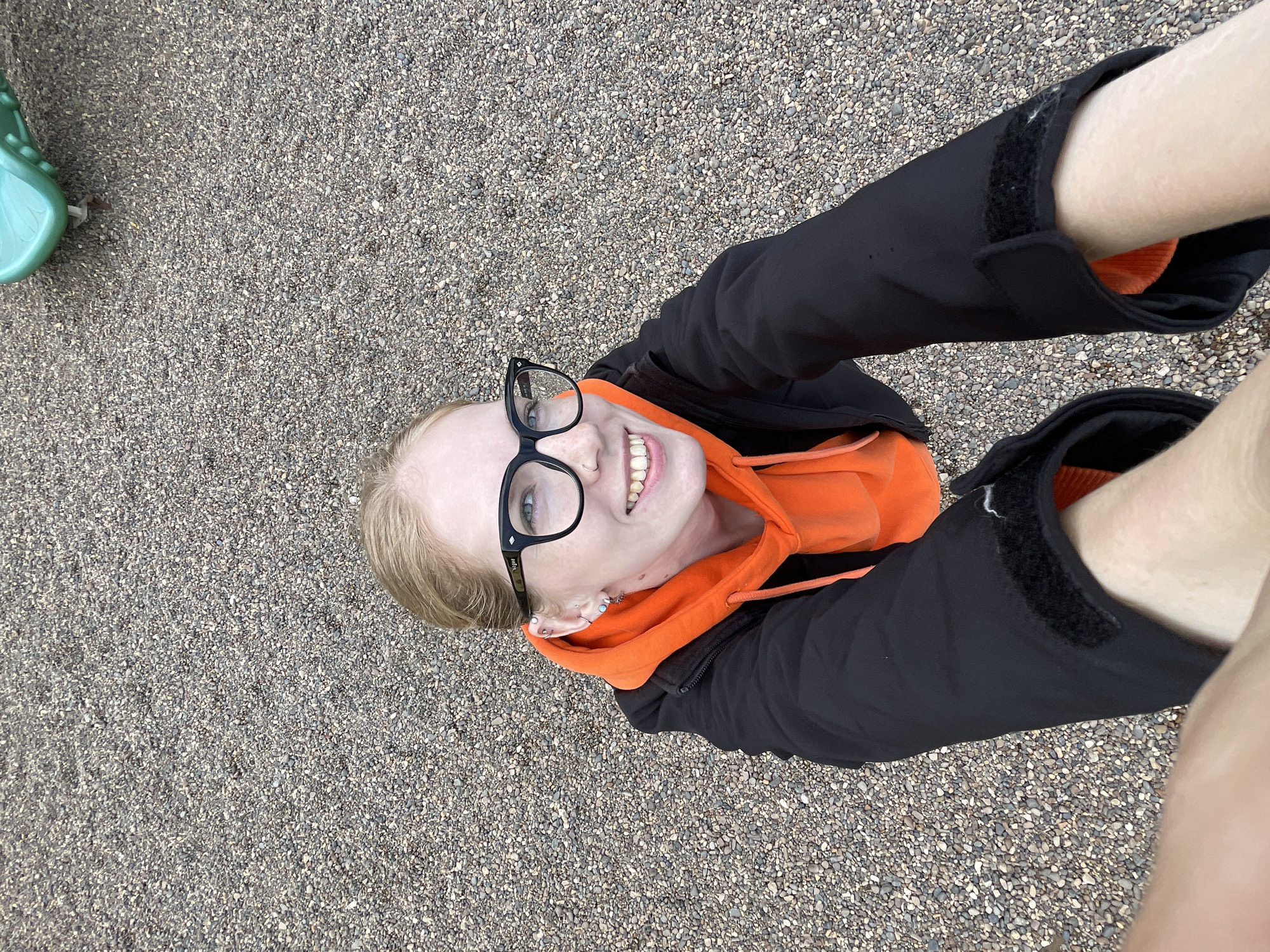 The image captures an overhead perspective of a young individual, likely female, smiling broadly while looking directly up at the camera. The ground beneath is covered in small, uniform grey and brown gravel. The subject has blonde hair and wears black-framed glasses, with the brand "Hype" faintly discernible on the left temple. Her blue eyes are bright, and her face, lightly freckled, displays a joyful expression with an open-mouthed smile revealing her teeth. She is dressed in a vivid orange hooded sweatshirt layered under a black zip-up jacket, which features Velcro closures on its cuffs. Her arms, clad in the black jacket, extend upwards towards the camera, while her lower legs or feet, appearing bare, enter the frame from the upper right, indicating a dynamic pose likely involving lying down, crouching, or sitting with limbs raised. A small, bright green plastic object is partially visible in the upper left corner, possibly part of playground equipment. The natural, bright illumination suggests a daytime capture. The overall feeling conveyed is one of happiness and carefree energy. This scene is located in Izhevsk, Russia.