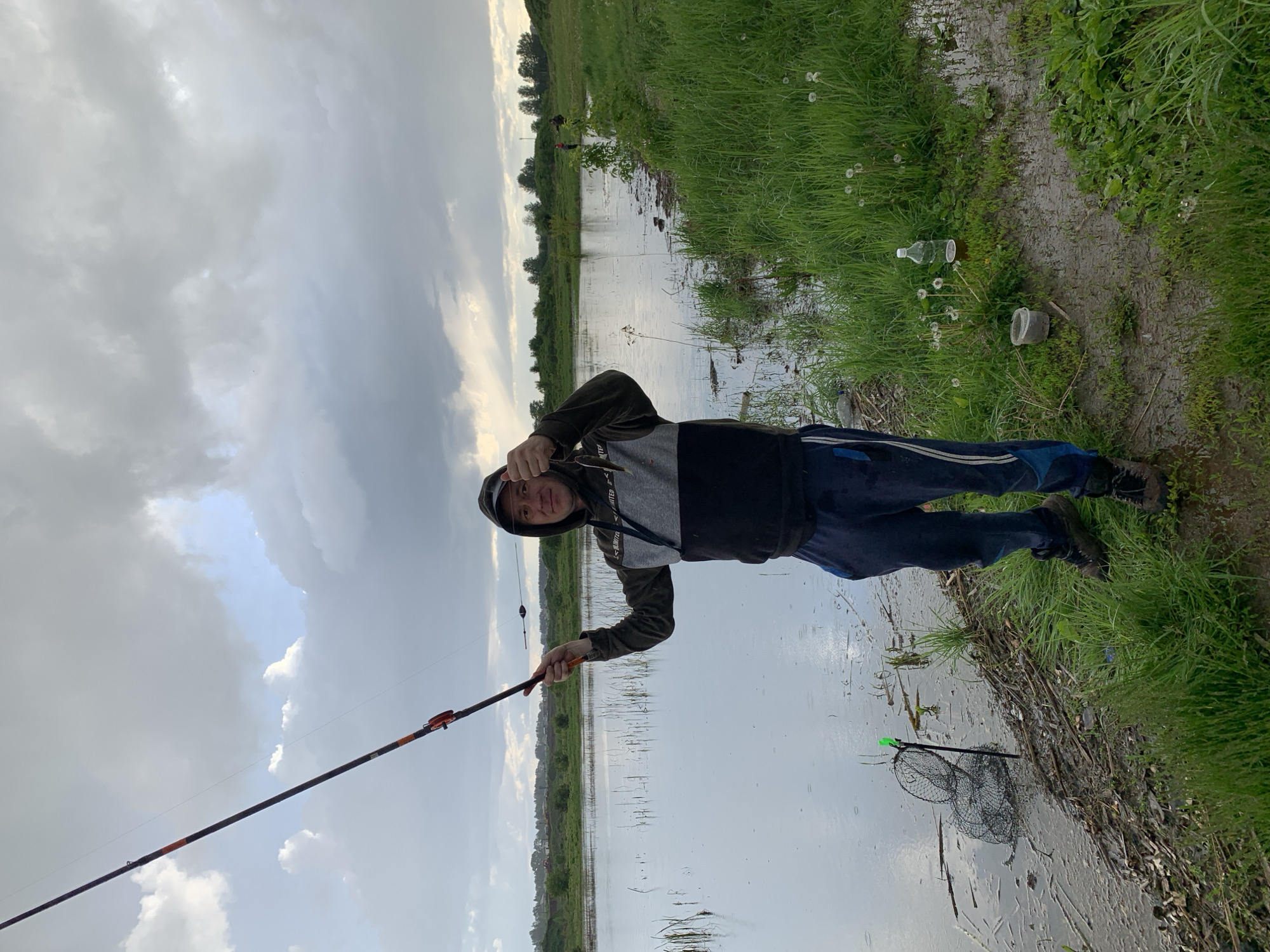 A man stands on the muddy bank of a body of water in Shaberdino, Russia. He is holding a small fish in his right hand and a fishing rod in his left. He wears a hooded sweatshirt, jeans, and dark shoes. A fishing net lies partially submerged in the water next to the bank. Trash, including plastic bottles and containers, litters the grass alongside the bank. The sky is overcast with gray and white clouds. The water appears calm, reflecting the sky. Grass and trees line the opposite bank.