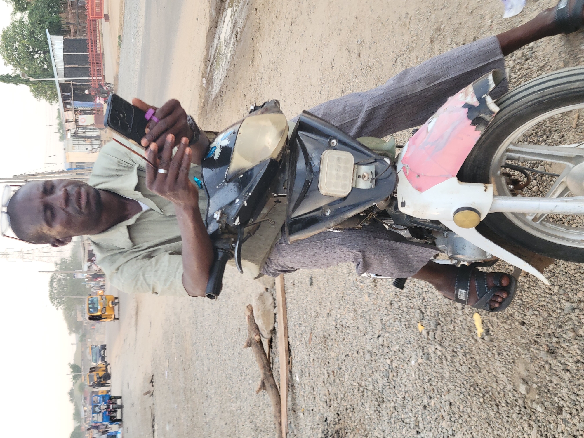 A man of African descent sits on a motorcycle on a partially paved road in Madalla, Nigeria. The man is dark-skinned and wears a light-colored collared shirt and gray trousers. He is holding a smartphone in his hands and appears to be looking at it. He wears a silver ring on his finger and is wearing black sandals.
The motorcycle is black and white with a faded pink section near the rear wheel. The motorcycle is parked on a road, which is made of both pavement and gravel.
In the background, yellow and blue vehicles are visible, along with buildings and trees. A wooden stick lays on the ground. The scene is set during daylight, with the lighting suggesting either early morning or late afternoon.