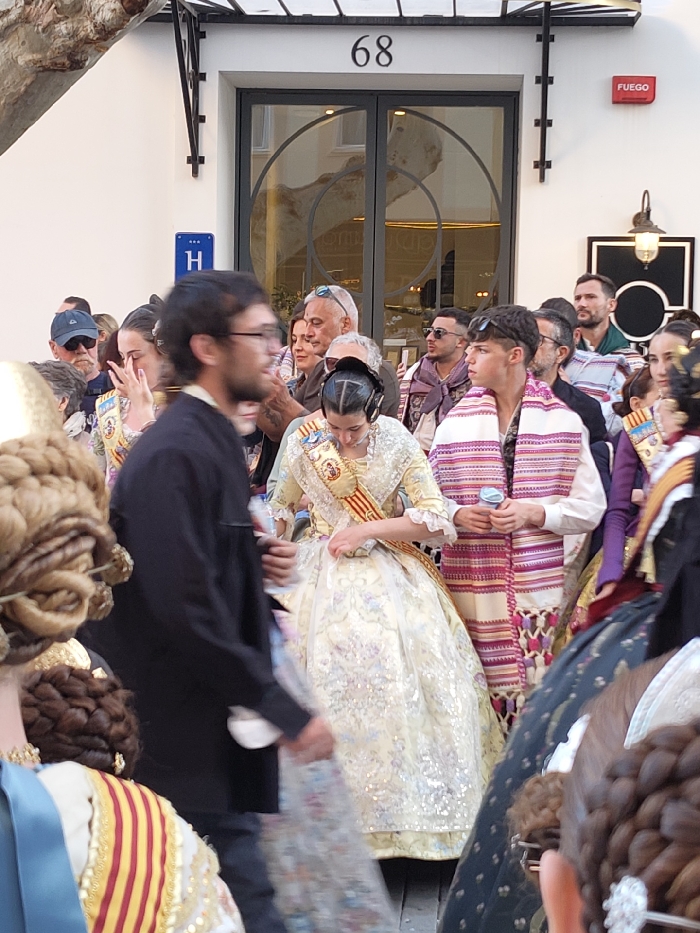 The scene depicts a crowded outdoor gathering in Dénia, Spain, in front of a cream-colored building numbered "68," identifiable as a hotel by a blue "H" sign. A red "FUEGO" sign is visible on the right wall.
In the midground, a young woman, centrally positioned, wears a traditional Valencian Fallera dress in pale yellow with intricate floral patterns and sequin embellishments. She has a sash displaying the Valencian flag colors with medals across her chest, and her hair is styled in a traditional Fallera bun, with black ear coverings (likely headphones or hairnets). She is looking downwards, appearing to adjust her dress. To her right, a young man wears a white shirt and a striped shawl in shades of purple, pink, and white with fringes. He holds a light blue object, possibly a microphone or small cup, and has sunglasses perched on his head.
In the blurred foreground, the back of two individuals' heads are visible on the left, displaying elaborate traditional Valencian hairstyles. A man with dark hair and glasses, wearing a dark shirt, walks from left to right, his form blurred from motion. The background features a diverse crowd of onlookers, including men and women of various ages, observing the central figures. The building features dark double doors with decorative circular glass inserts and dark metalwork extending above the entrance. A wall-mounted lantern is visible to the right of the doors.