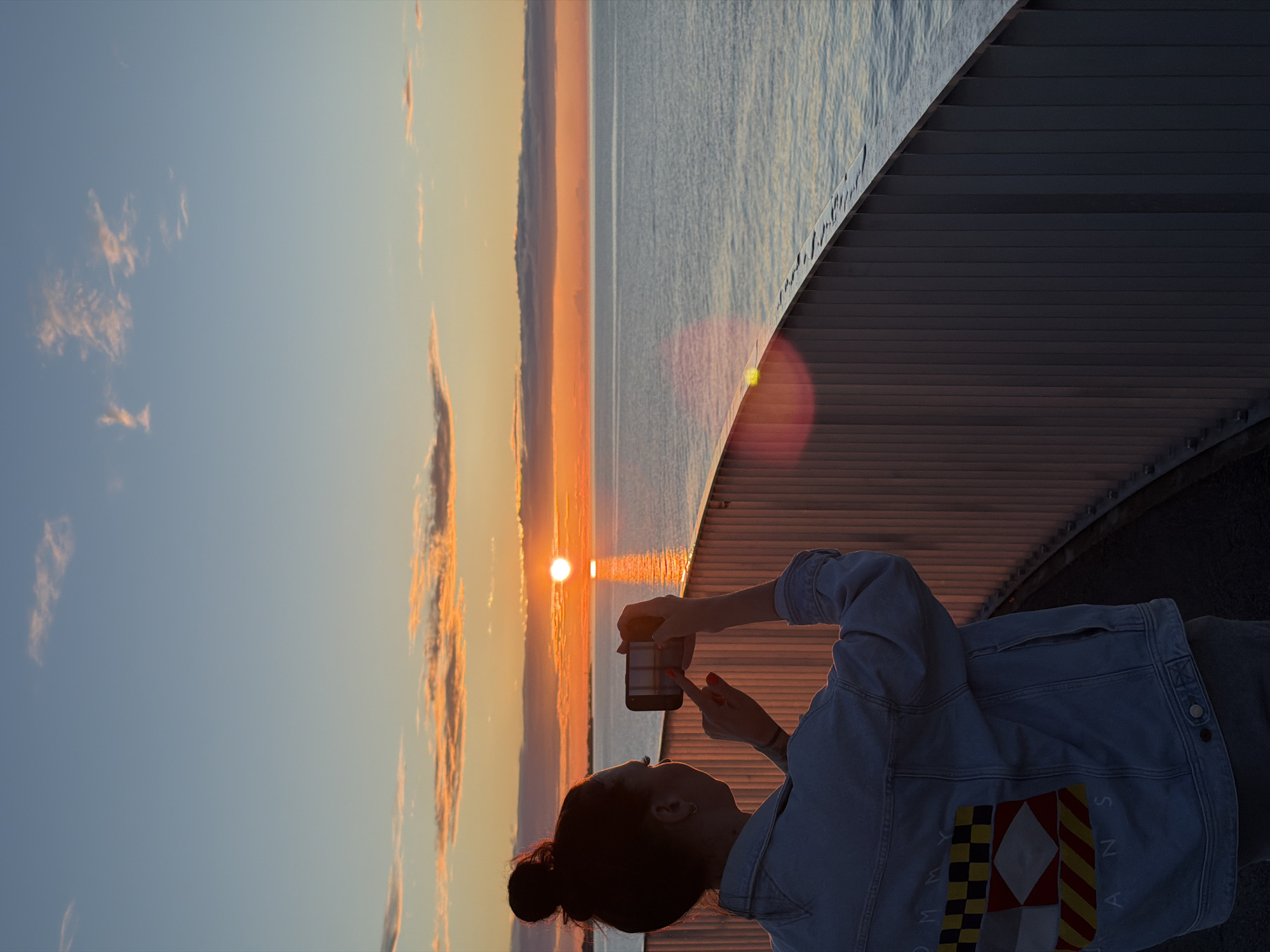 A woman, seen from behind, stands on a walkway at sunrise, taking a photograph of the sun with her smartphone. She has her hair pulled up in a bun and is wearing a denim jacket with a graphic design on the back. The sun is low on the horizon, casting a warm glow over the calm sea. The sky is a gradient of blue and orange, with wispy clouds. A railing separates the walkway from the sea. The location is the Pirita Promenade in Tallinn, Estonia, known for its scenic views of the Baltic Sea and the city skyline.