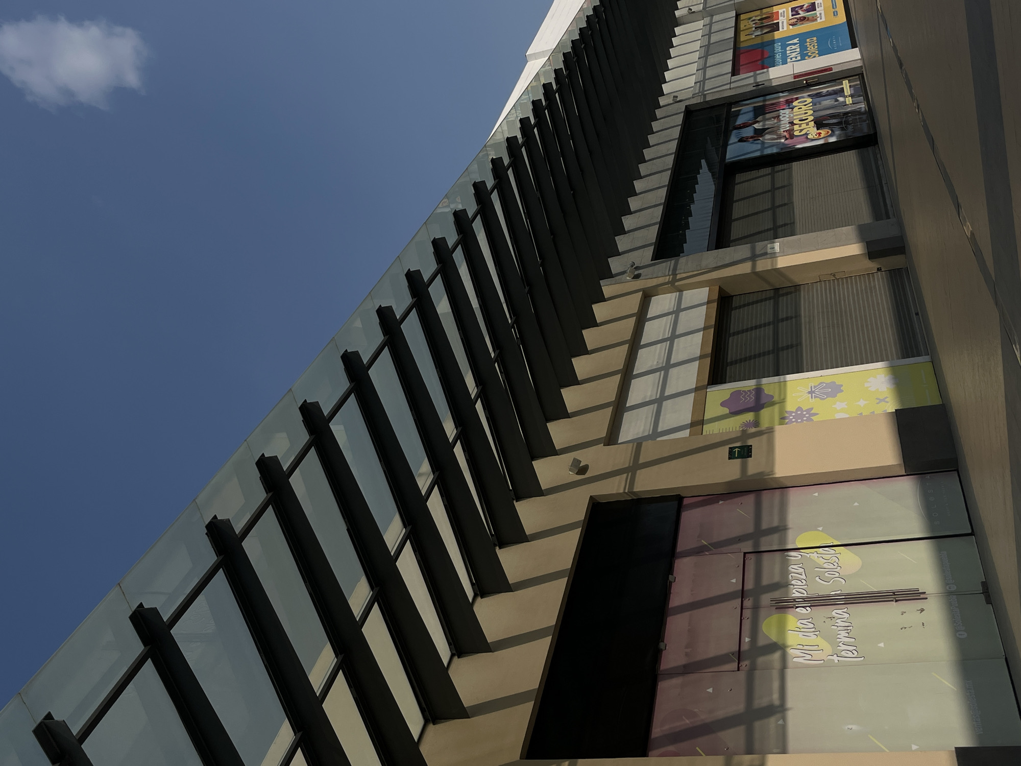 The image captures a modern building with a predominantly glass facade, punctuated by strong vertical black beams. The building is bathed in sunlight, creating sharp shadows that stretch across the concrete ledges, adding depth to the architectural features. The lower portion of the building reveals a ground-level storefront with commercial signage. Above, posters and advertisements are visible, hinting at the location being a commercial district. The sky is a clear, bright blue with a single, small white cloud in the upper left corner. Text in Spanish is present on the storefronts. The image appears to be taken from a low angle, emphasizing the height and vertical lines of the building. The angle is askew, with the building rotated slightly counterclockwise.