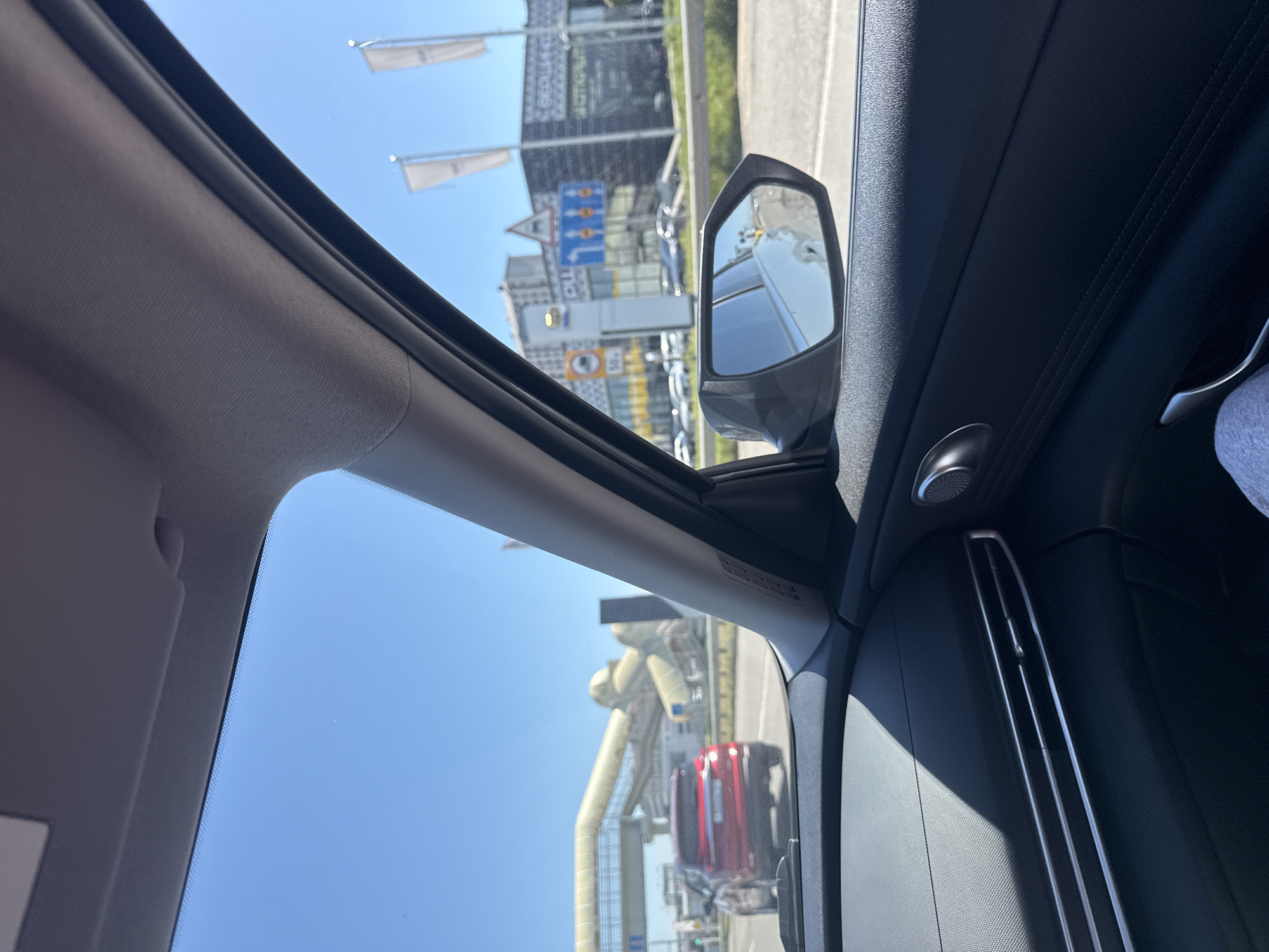 The image shows a view from inside a car, looking out the window. Outside, there is a road with a red truck visible. A building complex is seen in the background, including several flagpoles with white flags. The sky is clear blue. A side mirror is visible. The car interior is black. The location is Unknown, Unknown.