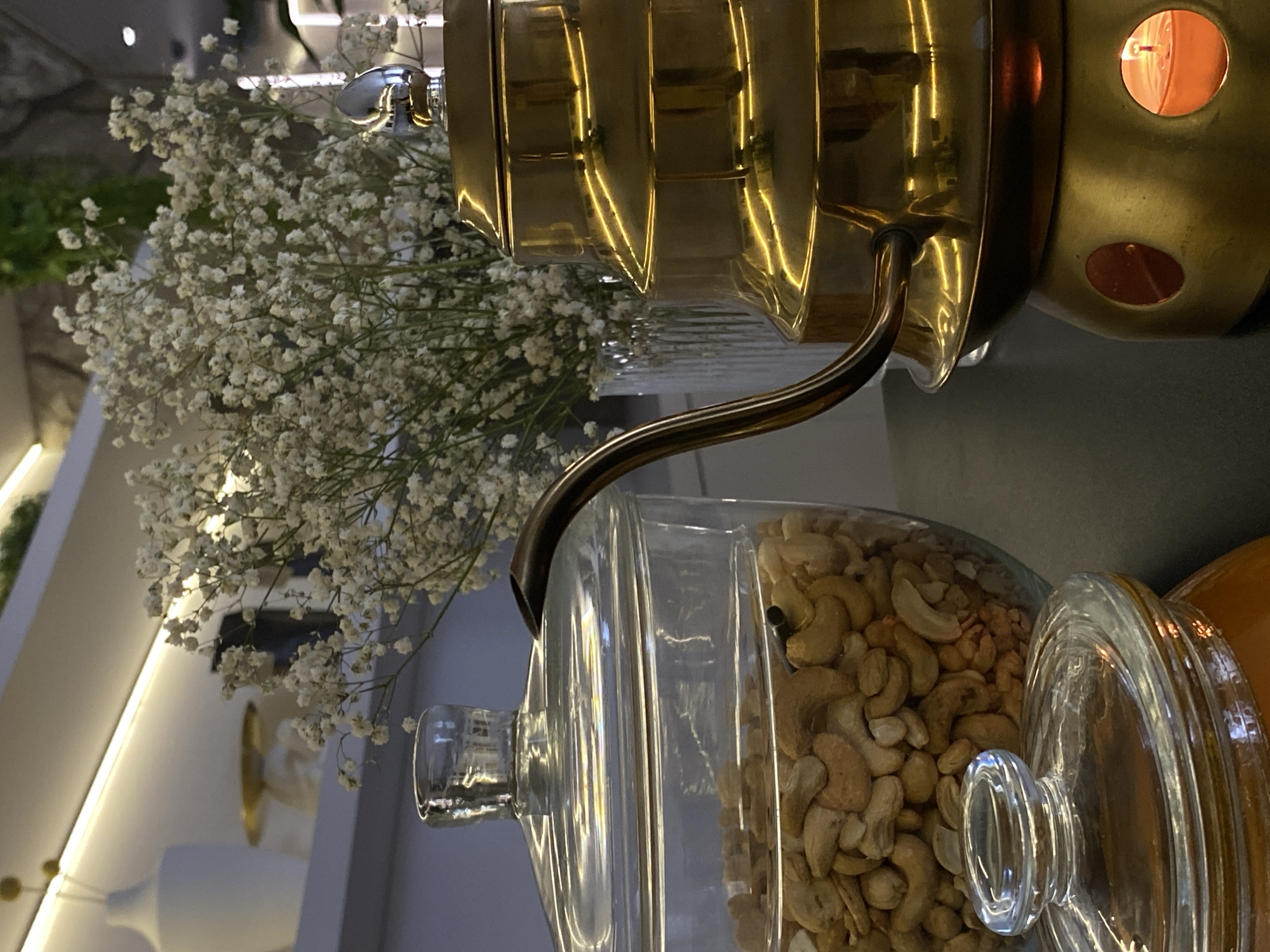 The image depicts a still life arrangement. A glass jar filled with cashews sits on a surface, likely a counter or table. To the left of the jar is a vase containing baby's breath flowers. Above the vase is a decorative brass or gold-colored item, possibly a lamp or container with a visible orange glow emanating from an opening.
The presence of the cashews suggests this may be a kitchen or dining area. The scene is indoors, illuminated by artificial light. The image does not reveal a specific location in Votkinsk, Russia, or any other city. There are no people visible in the frame.