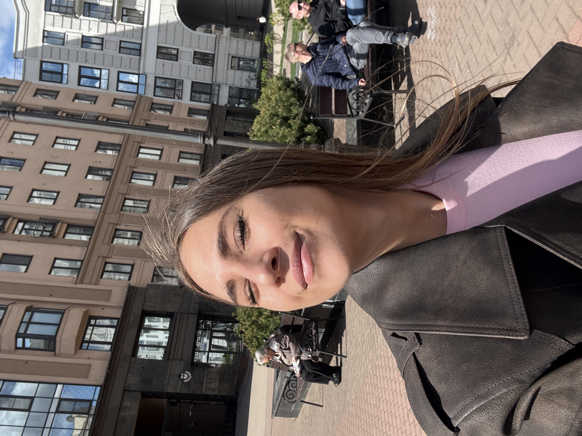 A person is taking a selfie outdoors. The person has light skin, brown hair, and is wearing a pink top and a black leather jacket. In the background, there are several buildings, some with ornate facades and many windows, suggesting an urban environment. People are sitting on benches, including one individual wearing a blue jacket. The brick-paved ground and architectural style suggest the location is likely within the city of Saint Petersburg, Russia.