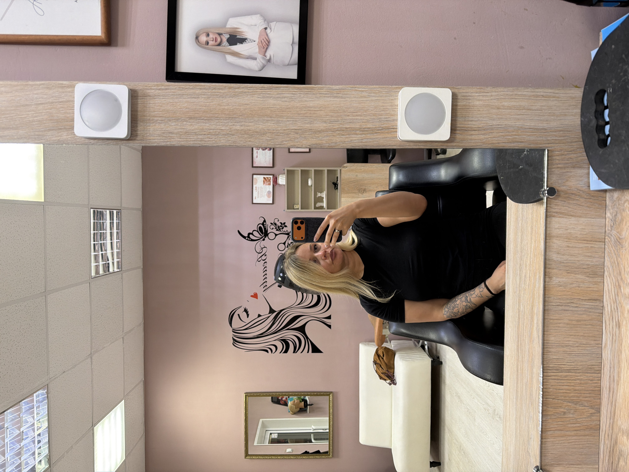 The scene is an interior shot in what appears to be a hair salon or beauty parlor. In the foreground, there's a large mirror framed with light wood. Within the mirror's reflection, a woman is taking a selfie with her phone. She has blonde hair and is wearing a black top. She is sitting in a black salon chair. A white cushioned chair is situated near the salon chair.
The wall behind the woman is a light pink color and decorated with a stylized graphic of a woman's face in profile with flowing hair intertwined with a pair of scissors and the word "Barber" written in cursive. There is also a framed mirror hanging on the wall.
Above the mirror frame, there are two framed images on the wall. Above those, there are two more pictures. There is also a small storage unit on the wall. The ceiling of the room has drop lighting.
The location of the scene is likely Sarapul, Russia, according to the request.