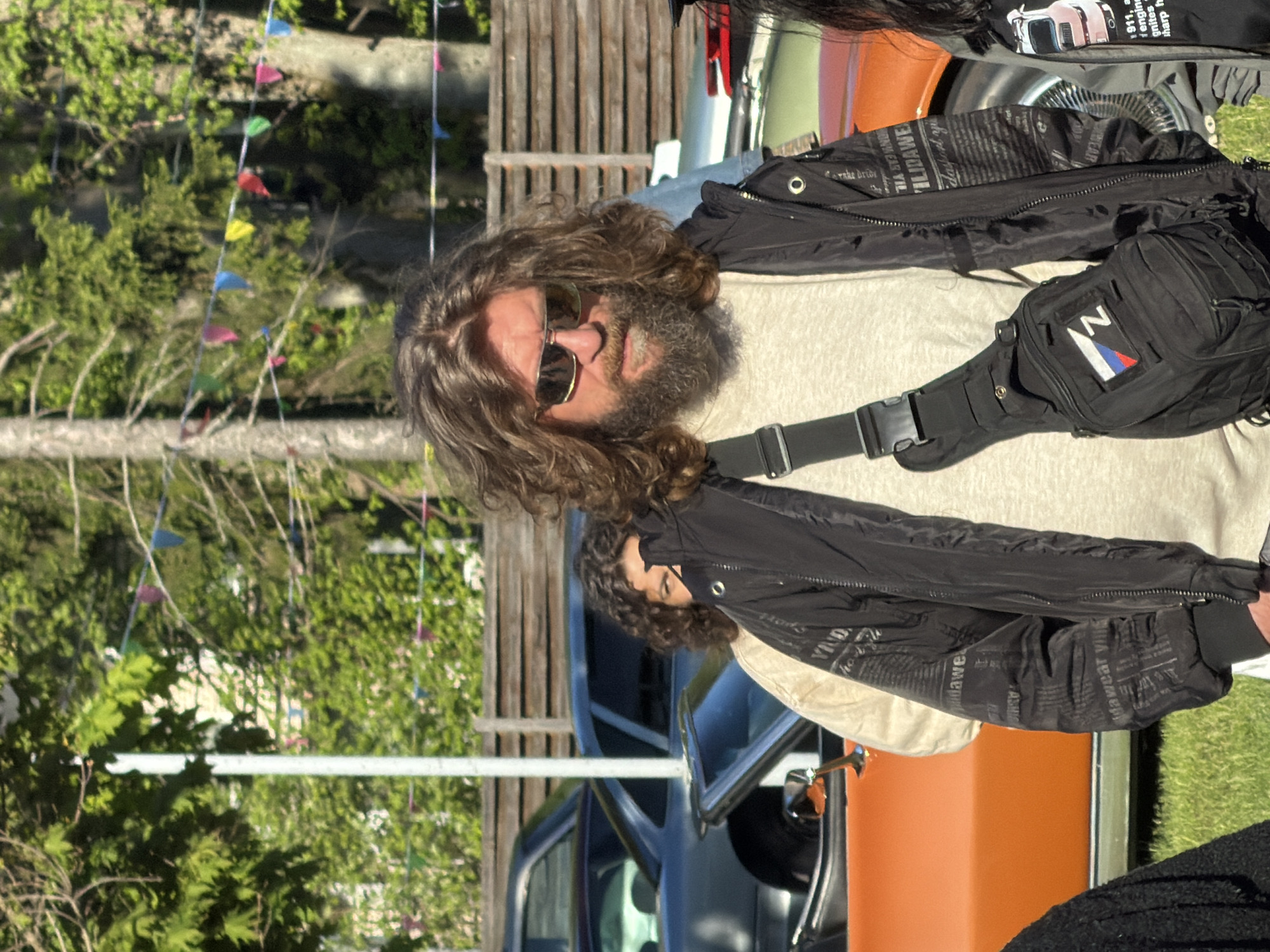 Here is a forensic description of the image:
**Subject:** The primary subject is a man with long, curly brown hair, a beard, and sunglasses. He is wearing a light-colored shirt, a black jacket with text prints, and a black fanny pack with a logo containing the colors red, blue, and white.
**People:** The image includes the man described above and a partial view of another person behind him with curly hair and a light-colored shirt.
**Objects:** Visible objects include trees, colorful triangular flags strung between trees, a wooden fence, two vintage cars (one orange and one grey), car tires, and the man's clothing accessories.
**Actions/Interactions:** The man is standing and looking towards the camera. The interaction between the two people is unclear, as only part of the second person is visible.
**Environment:** The scene appears to be outdoors, likely at a car show or event in a park-like setting. The background shows trees and decorative flags, suggesting a celebratory or festive atmosphere. There is grass.