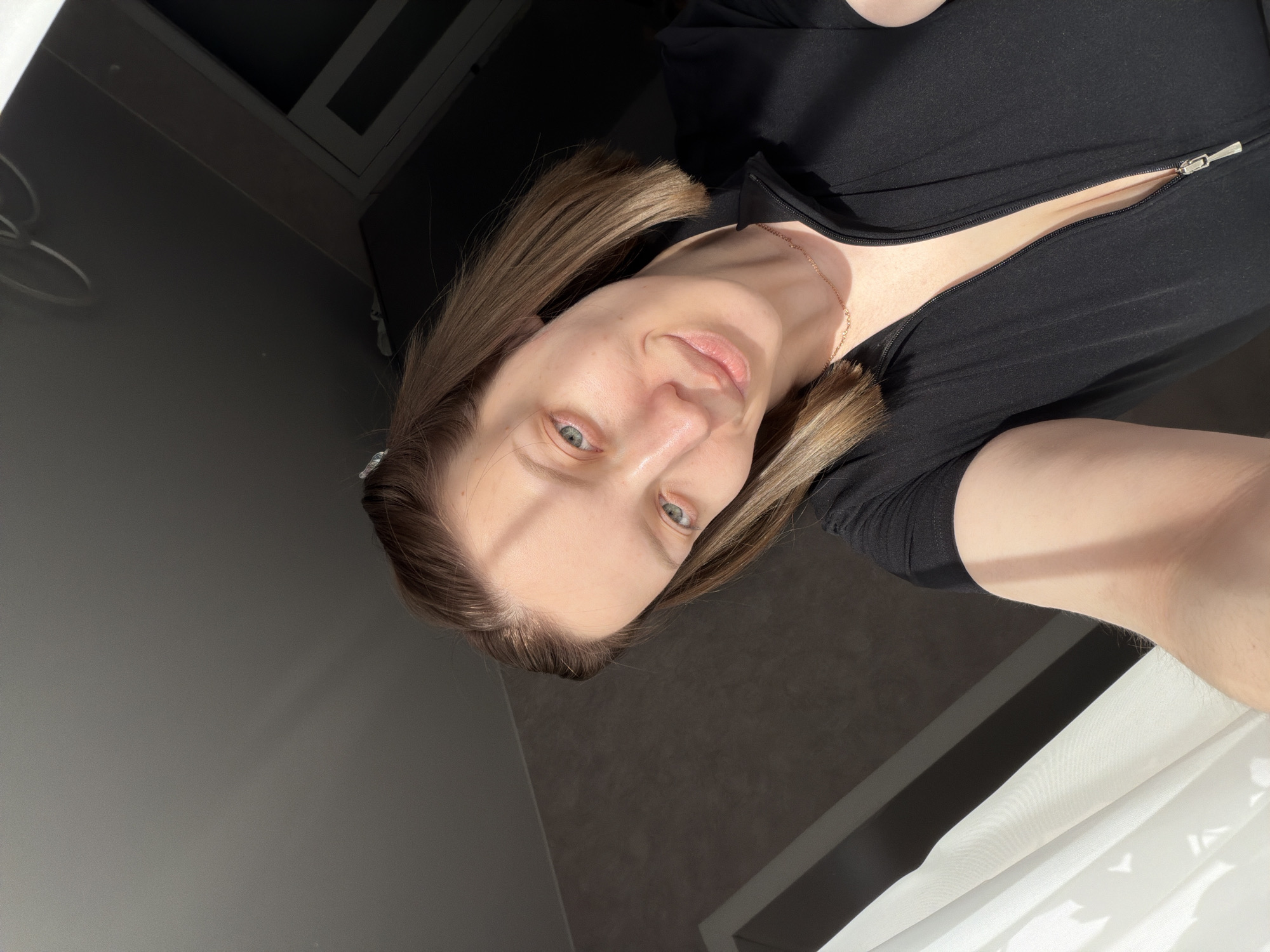 The image captures a selfie portrait of a woman, oriented with her head to the left and body extending to the right. She has light brown, shoulder-length hair and fair skin, with light-colored eyes (blue/green). Her expression is neutral with a slight hint of a smile as she looks directly at the camera. She is wearing a black top with a silver zipper that is partially open, revealing a glimpse of her chest. A thin, delicate gold chain necklace is visible around her neck.
The setting appears to be indoors, with natural light strongly illuminating her face and the white curtain on the right side of the frame, suggesting a window nearby. Shadows are prominent, indicating bright, direct sunlight. The background consists of dark grey or black walls, with some areas showing a slightly lighter grey. On the far right, a section of a white curtain is visible, featuring distinct patterns of light and shadow, possibly from objects outside the window or the window frame itself. A dark ceiling or upper wall area with a white frame is also visible in the top left corner. The activity is the woman taking a selfie. There is no visible text in the image. The location context for this scene is Reshetnikovo, Russia.