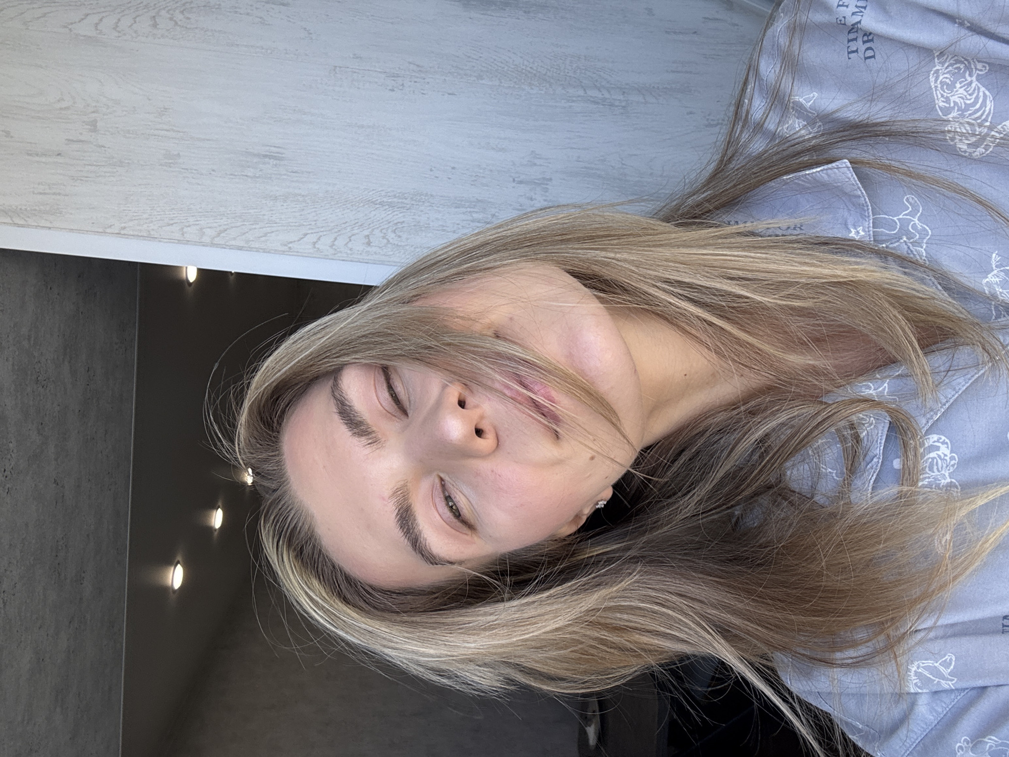 A young woman is lying down indoors, wearing a light blue pajama top with cartoon animal prints. Her head is angled towards the viewer, with closed eyes and a slight smile. She has light brown hair with blonde highlights, and visible eyebrows. Her face is well-lit. The background shows a gray wall on the left with three small, elongated lights, and a white textured wall above. The location appears to be inside a building in an unknown city.