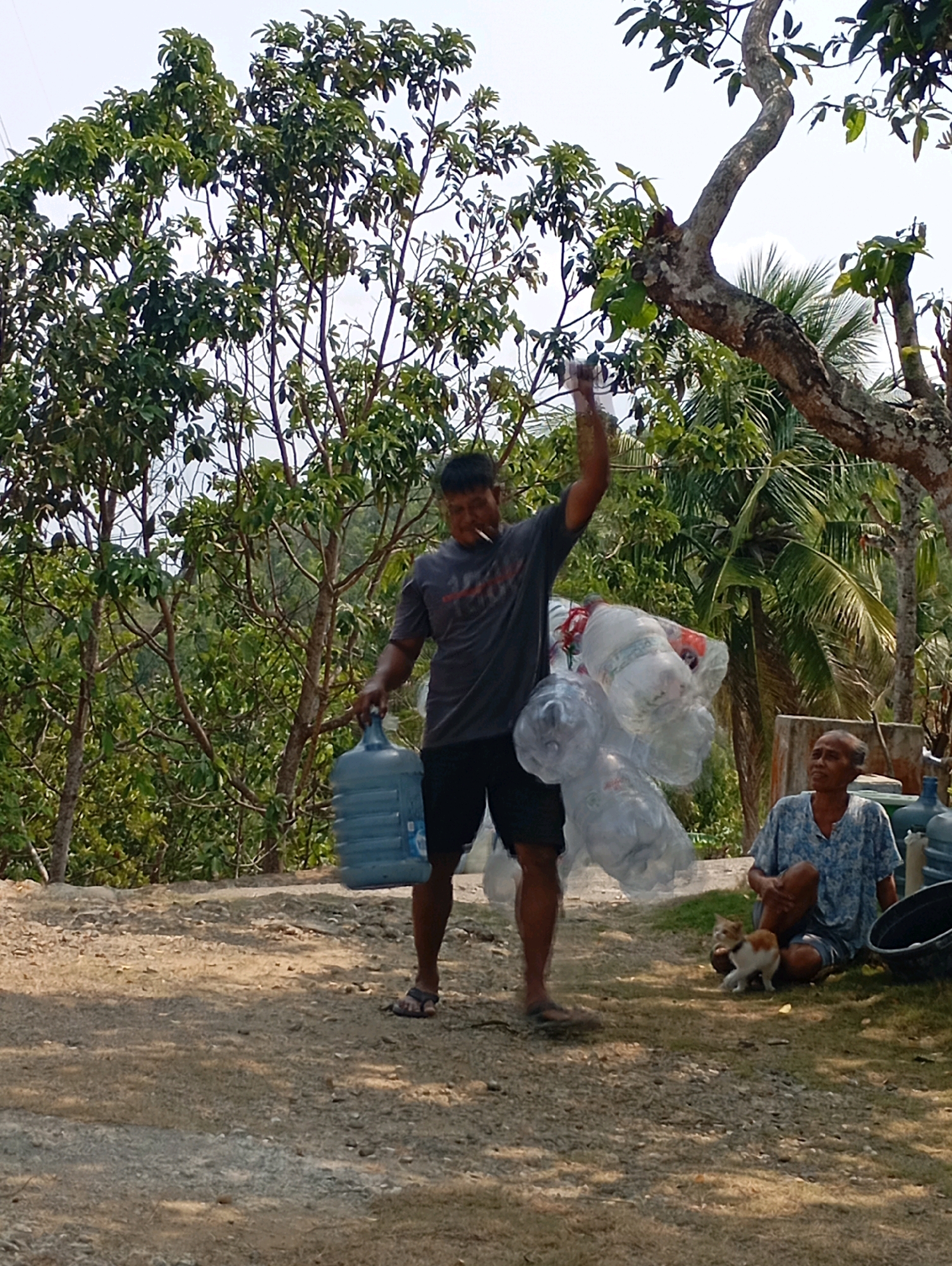 In Ketepung, Indonesia, a man wearing a gray t-shirt and black shorts is walking, carrying a large blue water jug in his left hand and a bundle of clear plastic water bottles tied together in his right hand. He appears to be smoking. To his right, an elderly woman is sitting on the ground, wearing a blue and white patterned shirt and dark shorts, looking to her left. A small, orange and white cat is walking in front of her. The ground is covered in gravel and dirt, with patches of green grass. In the background, there are lush green trees and foliage, with a bright sky overhead.
