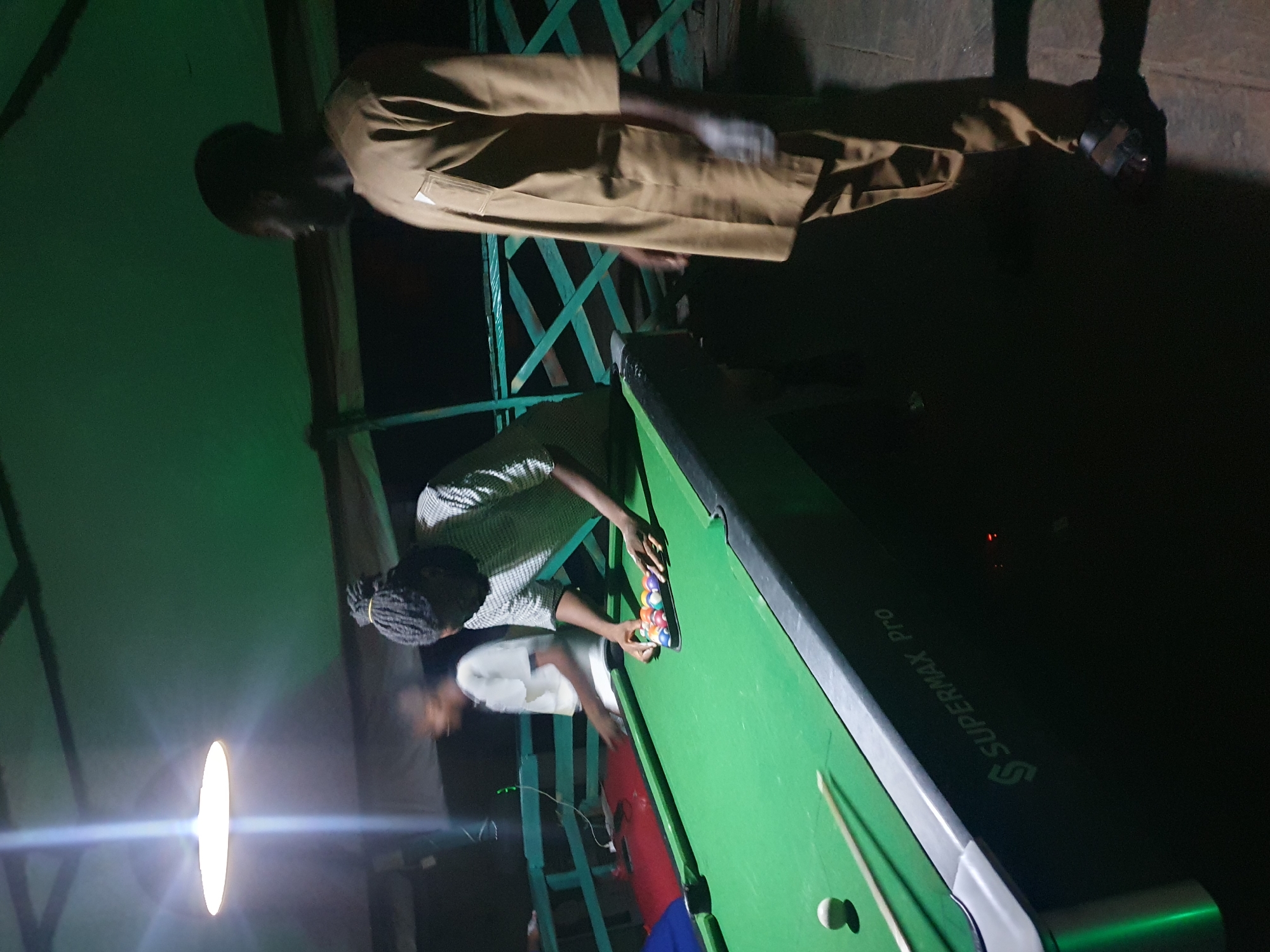 In Lokoja, Nigeria, three individuals are present at a billiard hall. Two men are actively engaged with a billiard table, with one man, wearing a patterned shirt, setting up a rack of billiard balls. Another man, in a tan robe, stands nearby, observing the game. A third person, wearing a white t-shirt, is blurred in the background, appearing to be walking away from the table. The billiard table itself is green and a cue stick and a single white ball are visible on its surface. The environment is dimly lit by an overhead lamp, and a green railing is visible in the background. The billiard table has "SUPERMAX Pro" branding visible on its side.