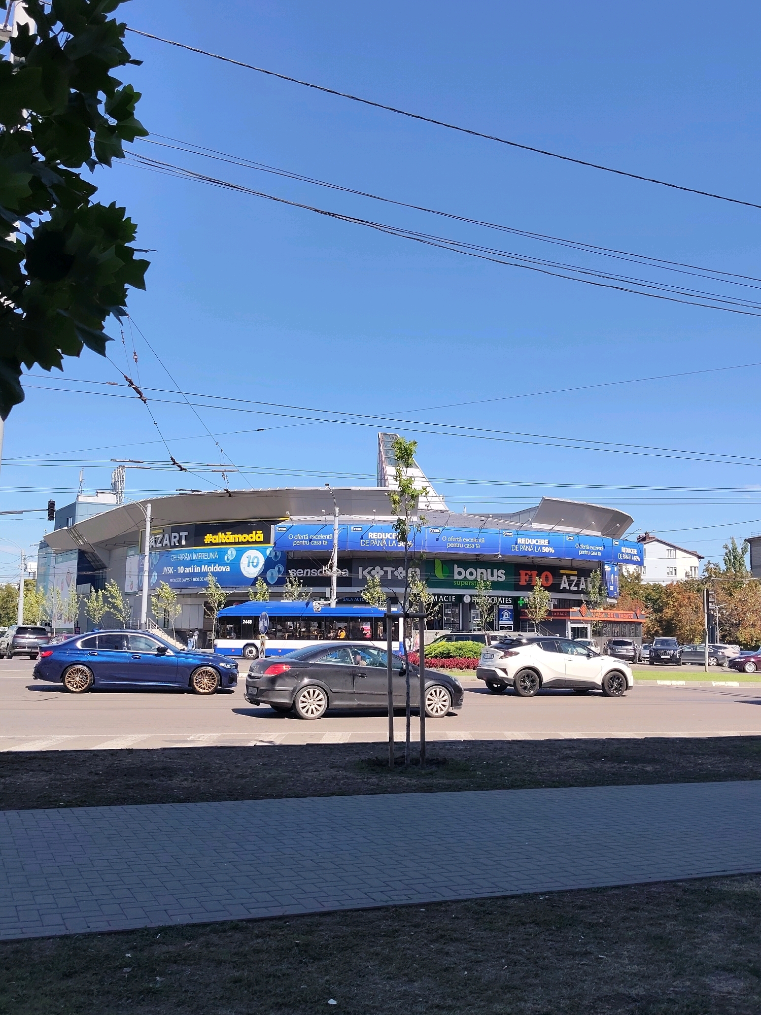 A street scene in an urban environment is captured under a clear blue sky. In the foreground, a paved pedestrian walkway borders a grassy area. Beyond the walkway, a paved road carries vehicular traffic. Several cars, including a blue sedan, a dark sedan, and a white crossover SUV, are visible on the road. A blue bus is also present. In the background, a large, modern building with a curved roof is adorned with various advertisements. Trees line the street, with a portion of a tree with dark green leaves in the upper left corner of the frame. Overhead, power lines crisscross the sky. The overall scene suggests a typical day in a commercial district.