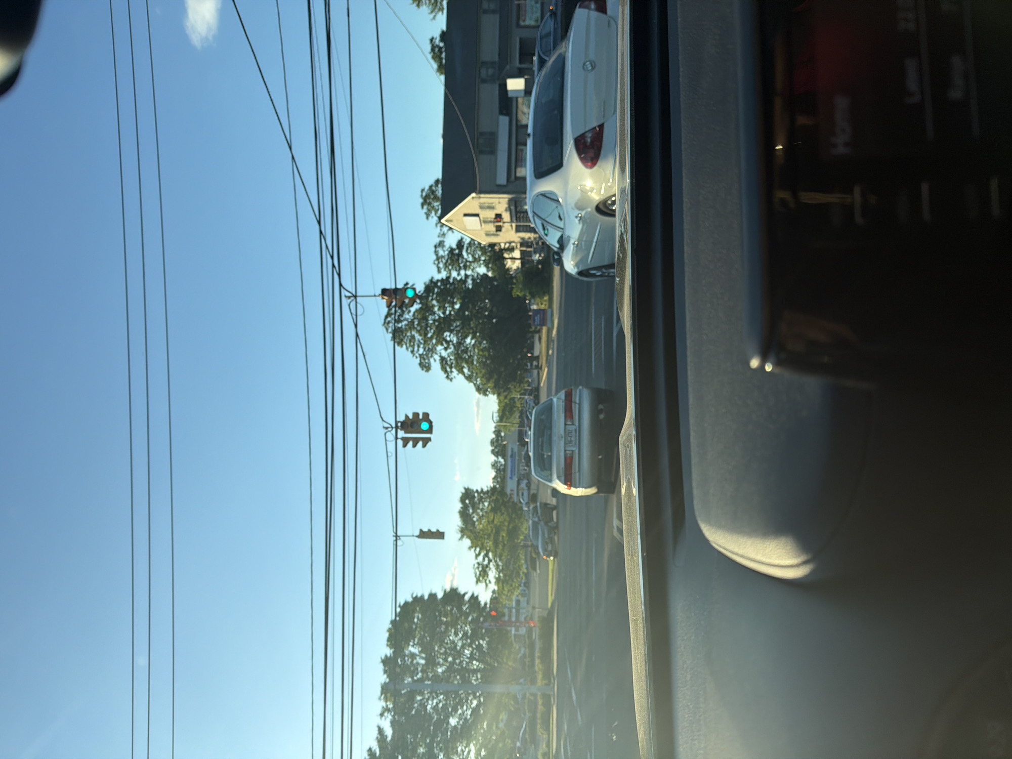 The image captures a view from the interior of a vehicle at a street intersection. The sky is a clear blue with a few faint clouds. Several power lines crisscross the upper portion of the frame.
In the foreground, the dashboard of the car is visible, with a portion of the entertainment system screen displaying the word "Home" and other text that is difficult to decipher.
Beyond the car, the intersection is active with traffic. Two traffic lights are illuminated with green signals. A silver sedan is stopped at the intersection, facing away from the camera. To the right of the silver sedan, a white sedan is visible, also at the intersection. Further in the background, several other vehicles and buildings line the street. Trees with green foliage are present on either side of the road, suggesting a suburban or commercial area. The sun appears to be low in the sky, casting long shadows and creating a warm light.
The scene is set in Riverhead, United States of America.