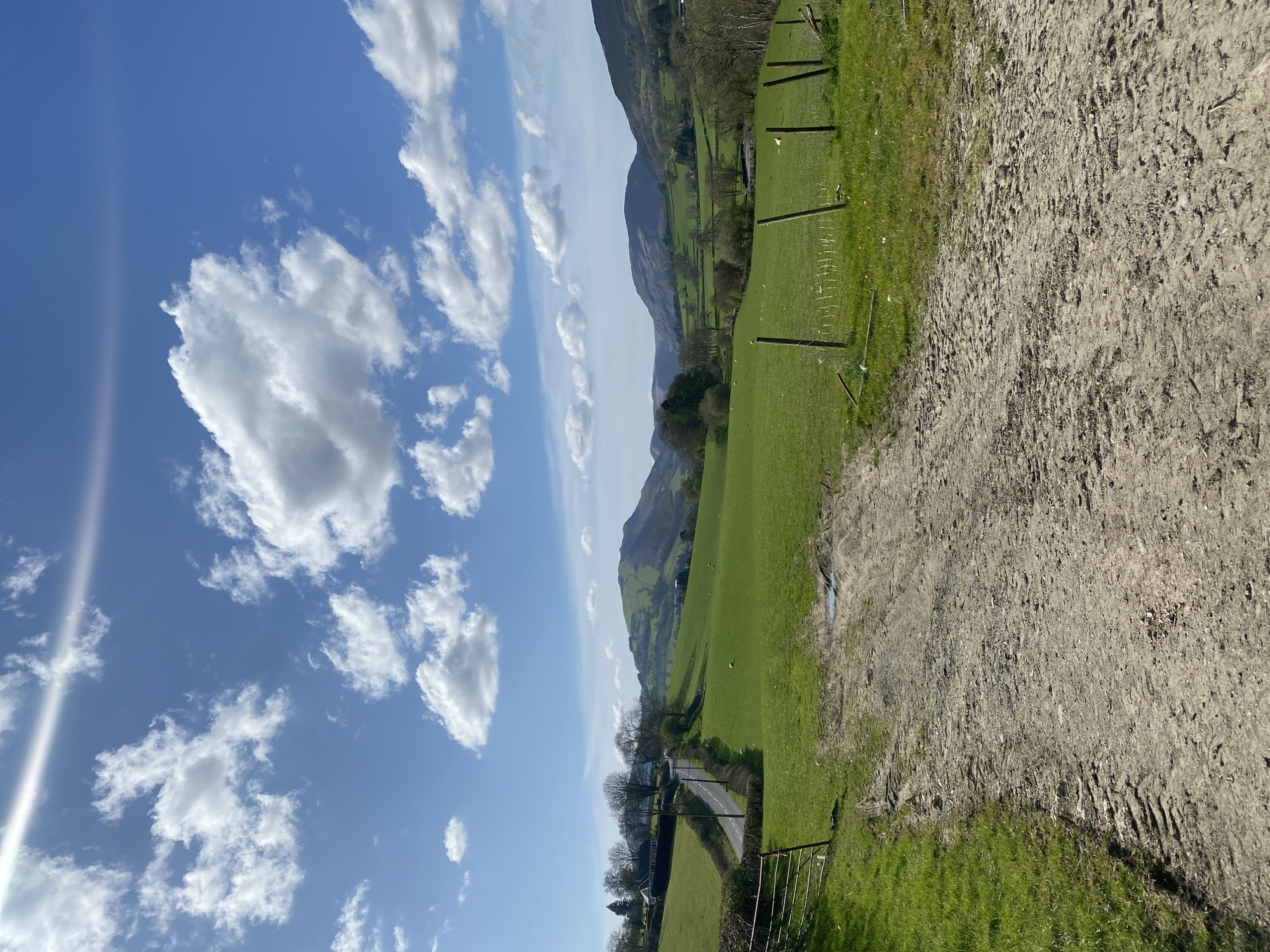 The image shows a rural landscape under a blue sky with scattered white clouds. A dirt path is visible in the foreground. Adjacent to the path is a green field, separated by a fence. Further into the background, a road lined with trees runs through the green landscape towards a mountain range. The location appears to be near Pen-y-bont-fawr, United Kingdom, based on typical characteristics of the area's landscape. There are no people visible. The primary subjects are the sky, fields, mountain, path and fences.