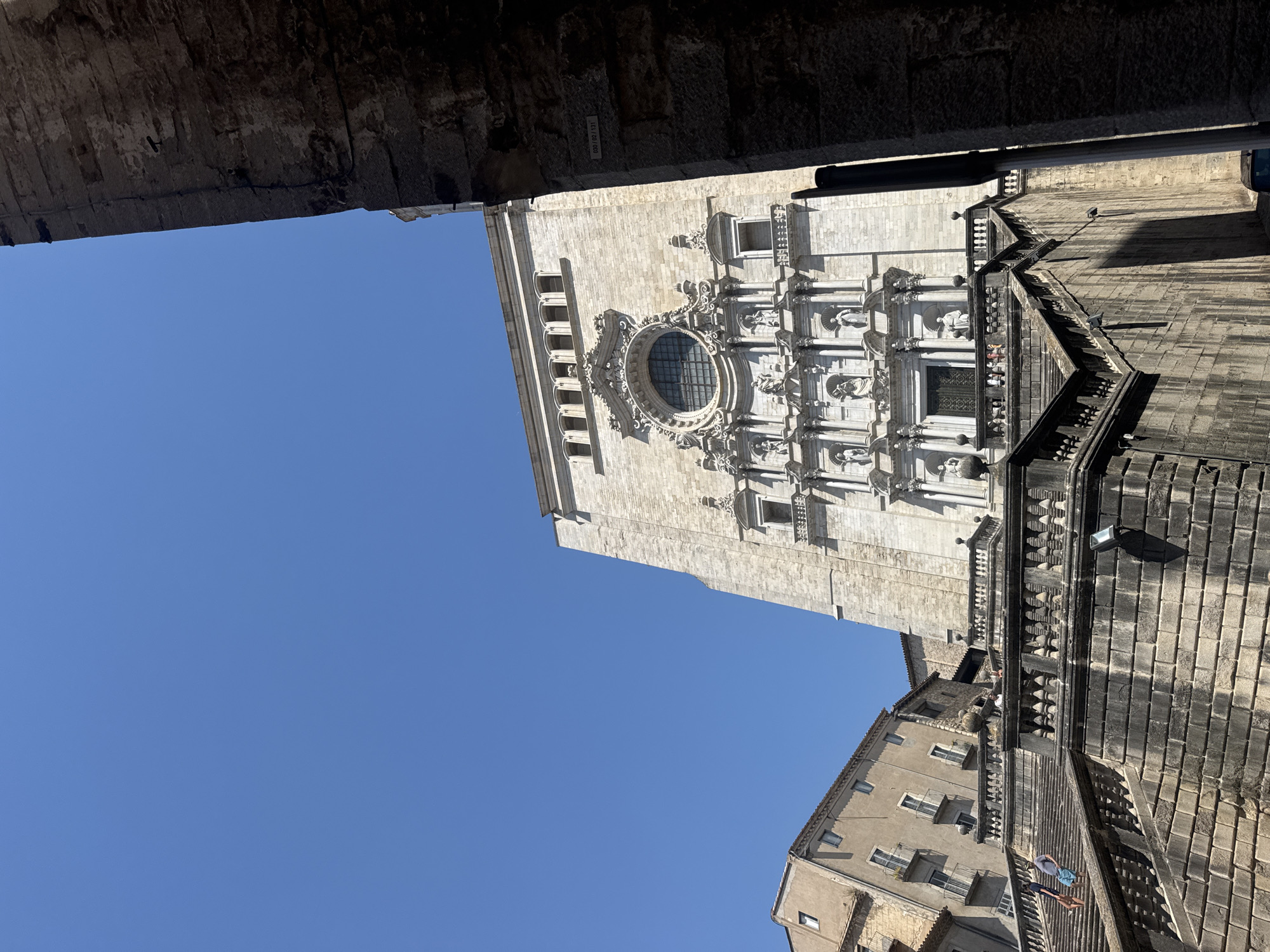 This is a low-angle, eye-level shot of the Girona Cathedral on a clear, sunny day. The cathedral's facade, made of pale stone, dominates the center of the frame, featuring intricate carvings, statues, and a large circular stained-glass window. In the foreground, a wide, stone staircase with ornate balustrades leads up to the cathedral's entrance. To the left of the cathedral, a building with a tan facade and several balconies is visible, its windows reflecting the bright blue sky. To the right, a dark, stone structure partially obscures the view of the cathedral. People can be seen walking up the stairs, interacting with the environment, and observing the architecture. The overall scene captures a majestic architectural landmark within a historic urban setting.