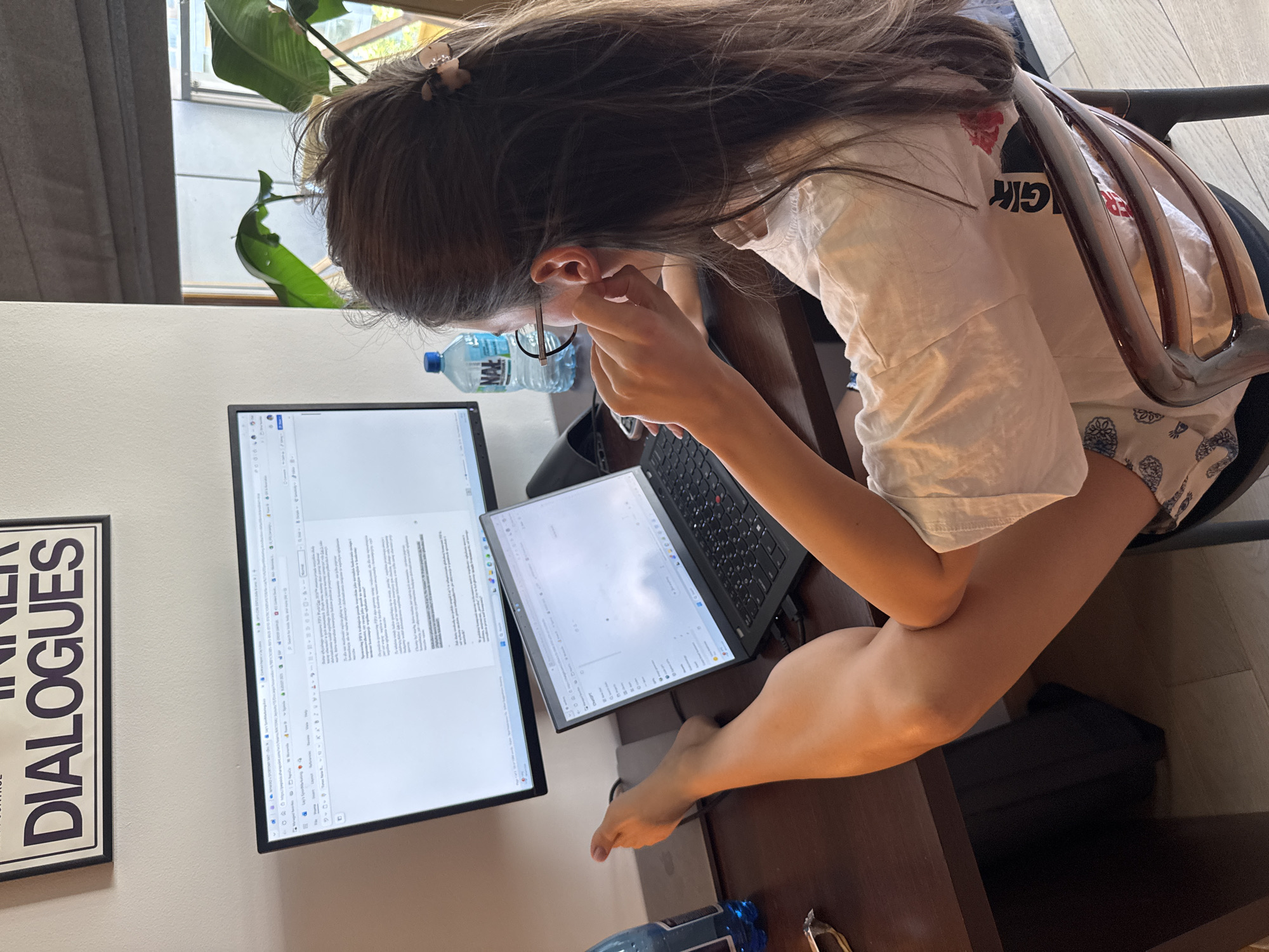 A woman is seated at a desk, working on a laptop and a larger monitor displaying a document, while also adjusting her glasses. A water bottle and a framed quote reading "INNER DIALOGUES" are visible on or near the desk. The location is indoors, but the specific city and country are unknown.