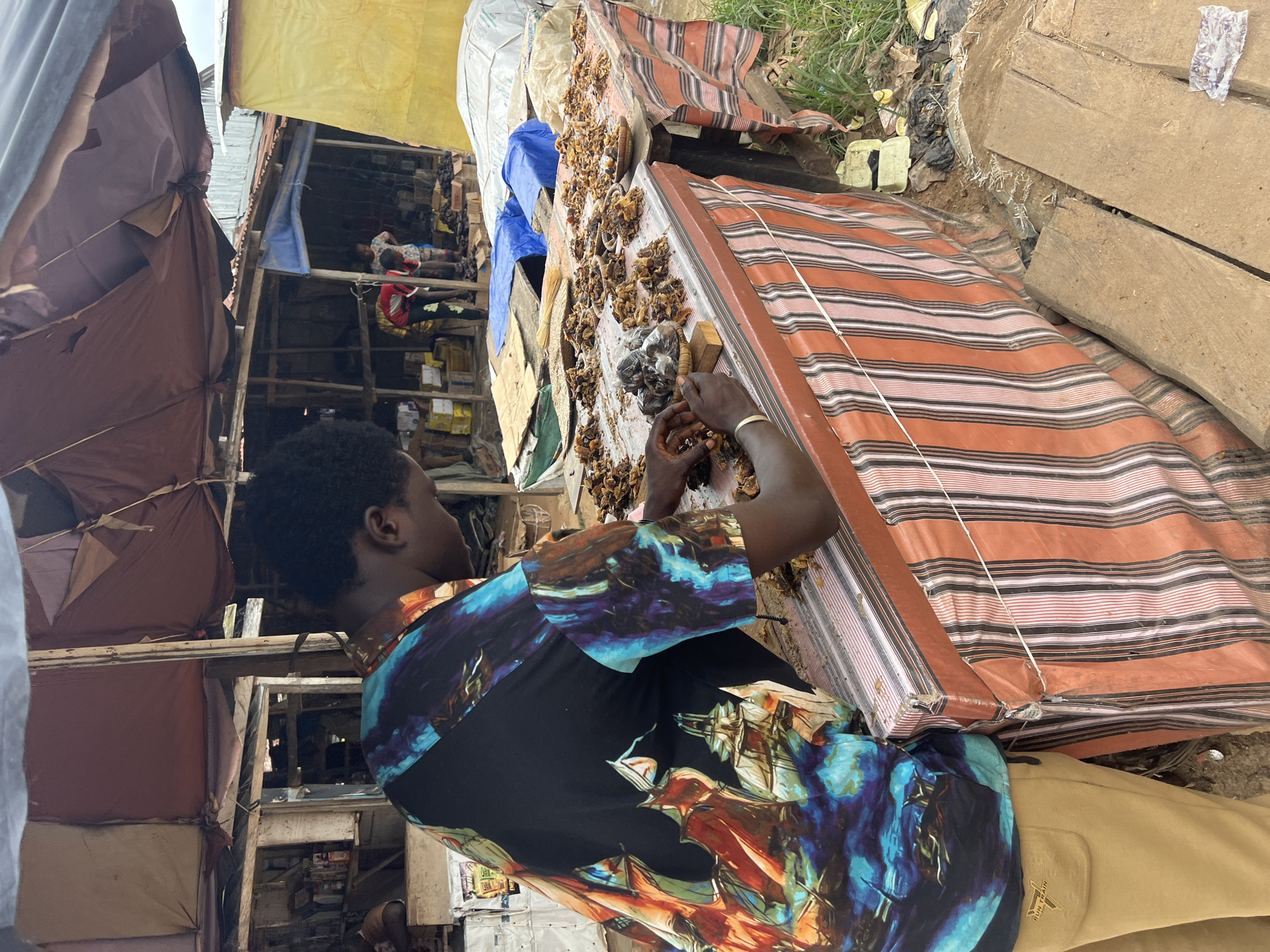 A person wearing a colorful, patterned shirt and tan pants is standing behind a table at an outdoor market in Asaba, Nigeria. The table is covered with a striped cloth and displays numerous dried food items, possibly fish or shrimp, arranged on plates and in small baskets. The person is handling some of the food items with their hands. The background reveals a makeshift market stall with a corrugated metal roof and wooden structures. Several other people are visible in the background, and there are various goods and packaging on display. The ground is dirt and debris, with some vegetation growing at the edge of the scene. The overall environment suggests a typical bustling open-air market.