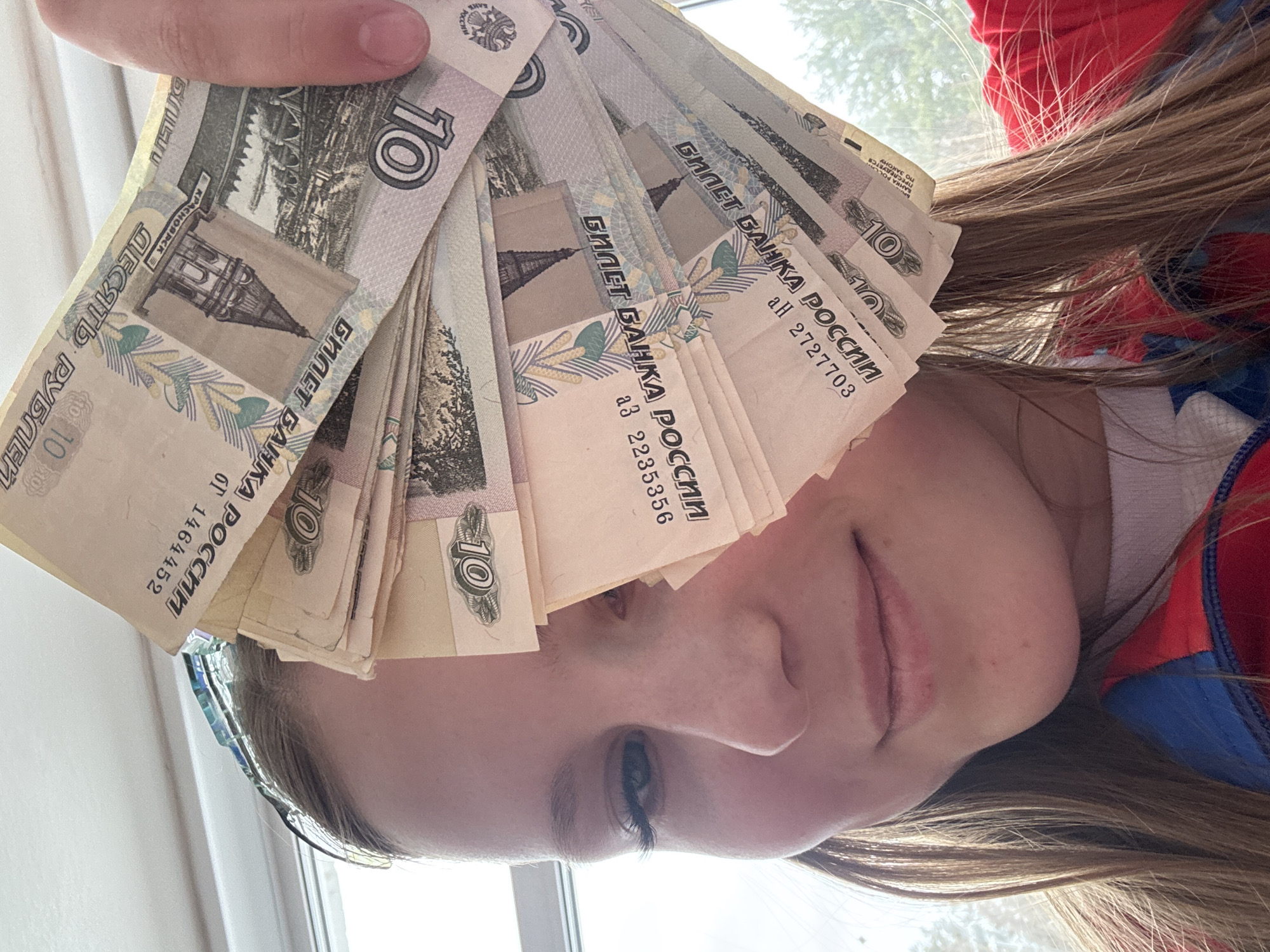 A selfie shows a person holding a stack of Russian 10 ruble banknotes in front of their face. The banknotes are predominantly light brown and depict architectural elements, specifically, the Krasnoyarsk Bridge, located in Krasnoyarsk. The person has light brown hair and is wearing a red and blue garment. A window is visible in the background, offering a glimpse of the outside. The photo appears to be taken indoors.