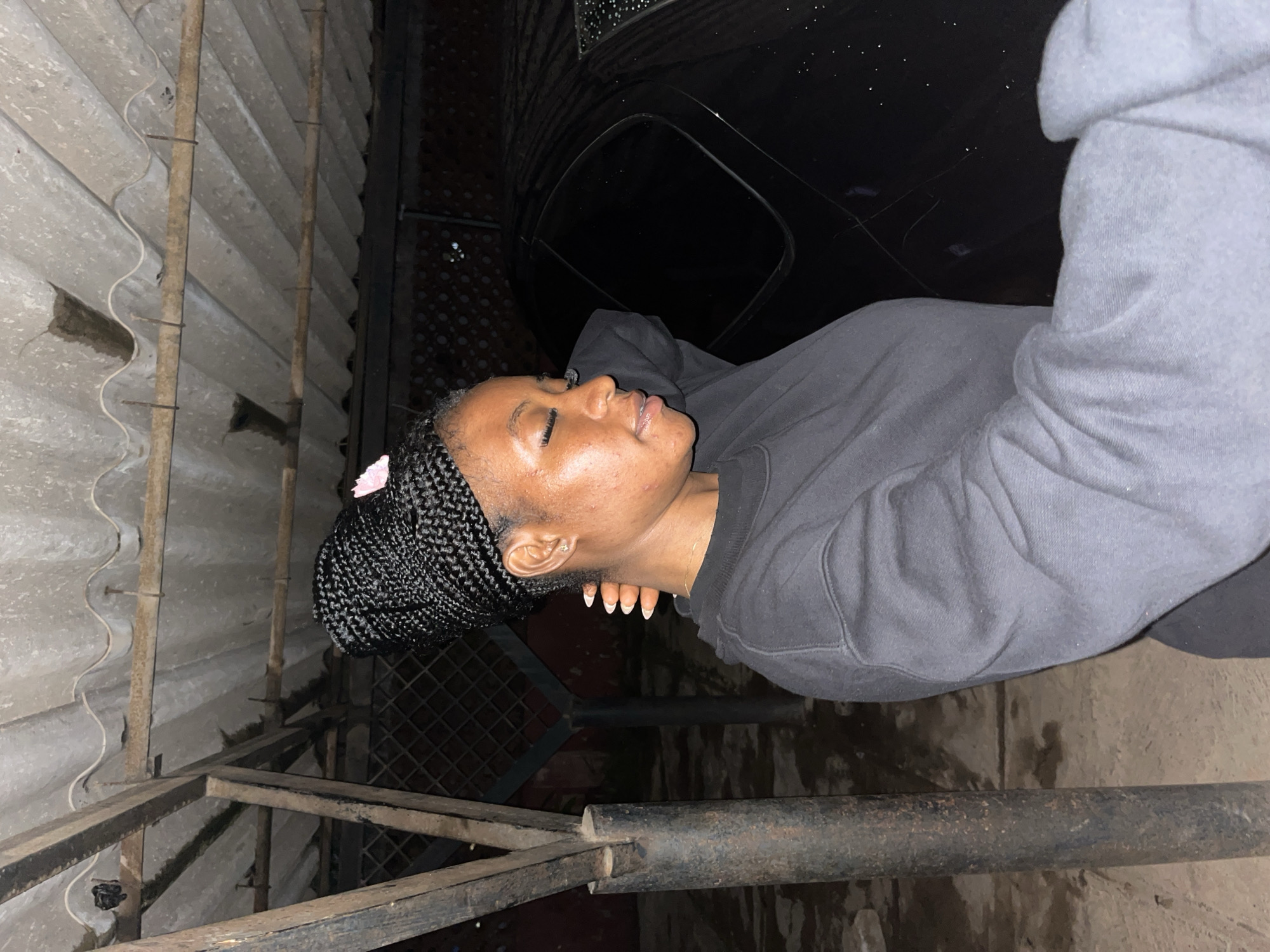 The image shows a young Black woman with braided hair standing in what appears to be an outdoor parking area under a corrugated metal roof. She is wearing a dark gray sweatshirt. The woman is positioned on the left side of the frame, with her head turned slightly to the right and her eyes closed. Her right hand is behind her head, and her left hand is extended forward, partially out of frame. A black car is visible to the right of the woman, and its surface appears wet with small water droplets. The ground is a light-colored, uneven surface, possibly concrete or packed dirt. The background includes some metal supports and a chain-link fence. The overall lighting suggests it is nighttime or dusk. Without further information, it is impossible to determine the specific location within Jos, Nigeria, other than it being an outdoor, likely residential or communal parking space.