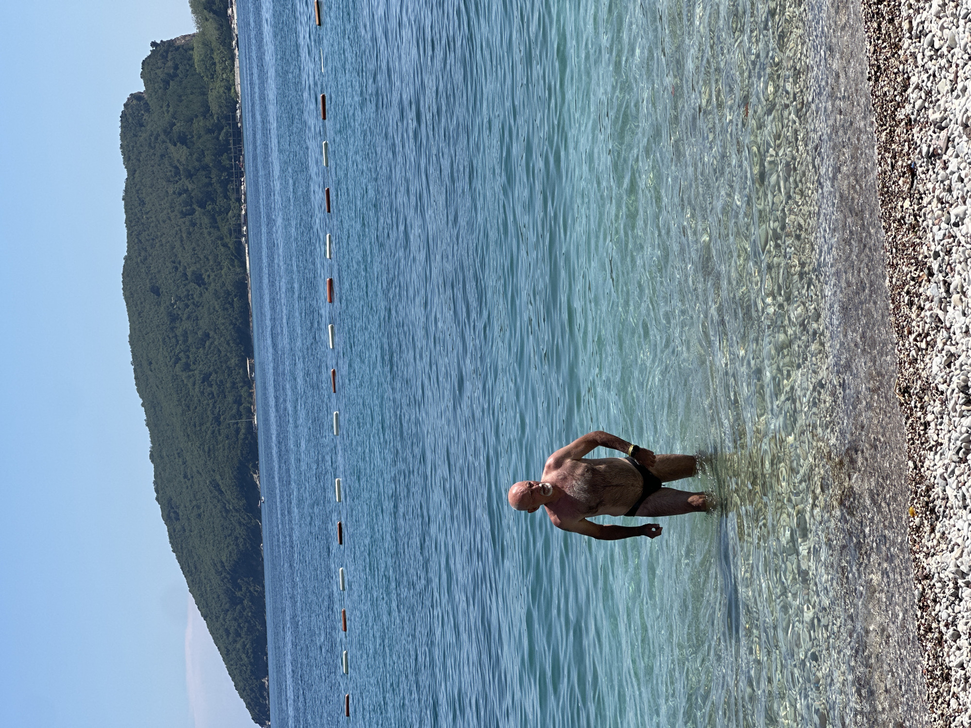 A man stands in the shallow, clear turquoise water of the Adriatic Sea at Mogren Beach, located west of the Old Town of Budva, Montenegro. The man is bald and has a light complexion, wearing dark swim briefs. His right hand is on his hip, and his left arm is bent at the elbow, with his hand resting on his thigh. The seabed is visible through the clear water, covered in small, light-colored pebbles. In the background, a line of buoys marks the swimming area, and beyond that, a large, tree-covered island rises from the sea. The sky is a clear, bright blue, indicating a sunny day. The beach in the foreground is also covered in pebbles.