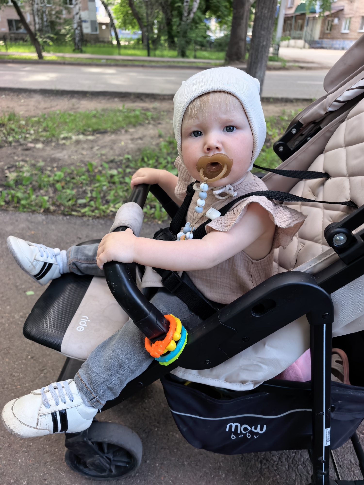 A young Caucasian child, approximately one to two years old, is seated in a stroller. The child wears a light-colored beanie, a pale pink textured dress with ruffles on the sleeves, gray denim jeans, and white sneakers with black stripes. The child has a pacifier in their mouth and a beaded pacifier clip attached to their dress. The stroller is beige and has black trim and wheels. A colorful toy ring is attached to the stroller's handle. The scene appears to be outdoors, possibly in a residential area of Sterlitamak, Russia, with a paved walkway or road in the foreground and a grassy area with trees and buildings in the background. The lighting suggests it is daytime.
