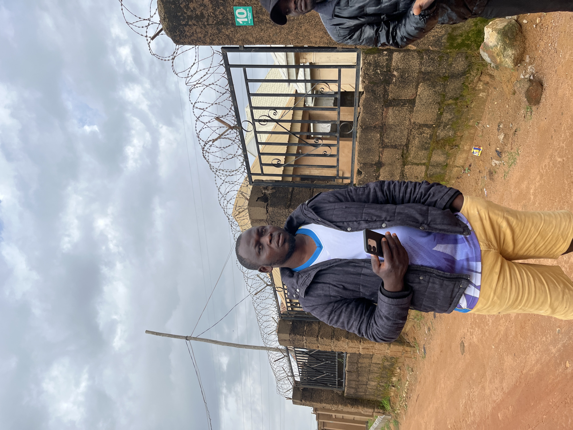 Two men stand outdoors in front of a brick wall topped with barbed wire. The man on the left is in the foreground, wearing a black puffer jacket over a white t-shirt with a blue and purple abstract design, and light yellow shorts. He holds a dark-colored smartphone in his right hand. His left hand is in his jacket pocket. He faces the camera with a neutral expression. The second man, partially visible on the right, wears a black puffer jacket and a black baseball cap. He is looking to his right. Behind them, a black metal gate with decorative scrollwork is set into the brick wall. Beyond the gate, houses with corrugated metal roofs are visible. A green sign with the number "10" is affixed to the wall near the gate. The ground in front of the wall is a reddish-brown dirt path. The sky above is overcast with gray clouds. A wooden pole with wires attached is visible to the left of the men. This scene appears to be in the city of Jos, Nigeria.