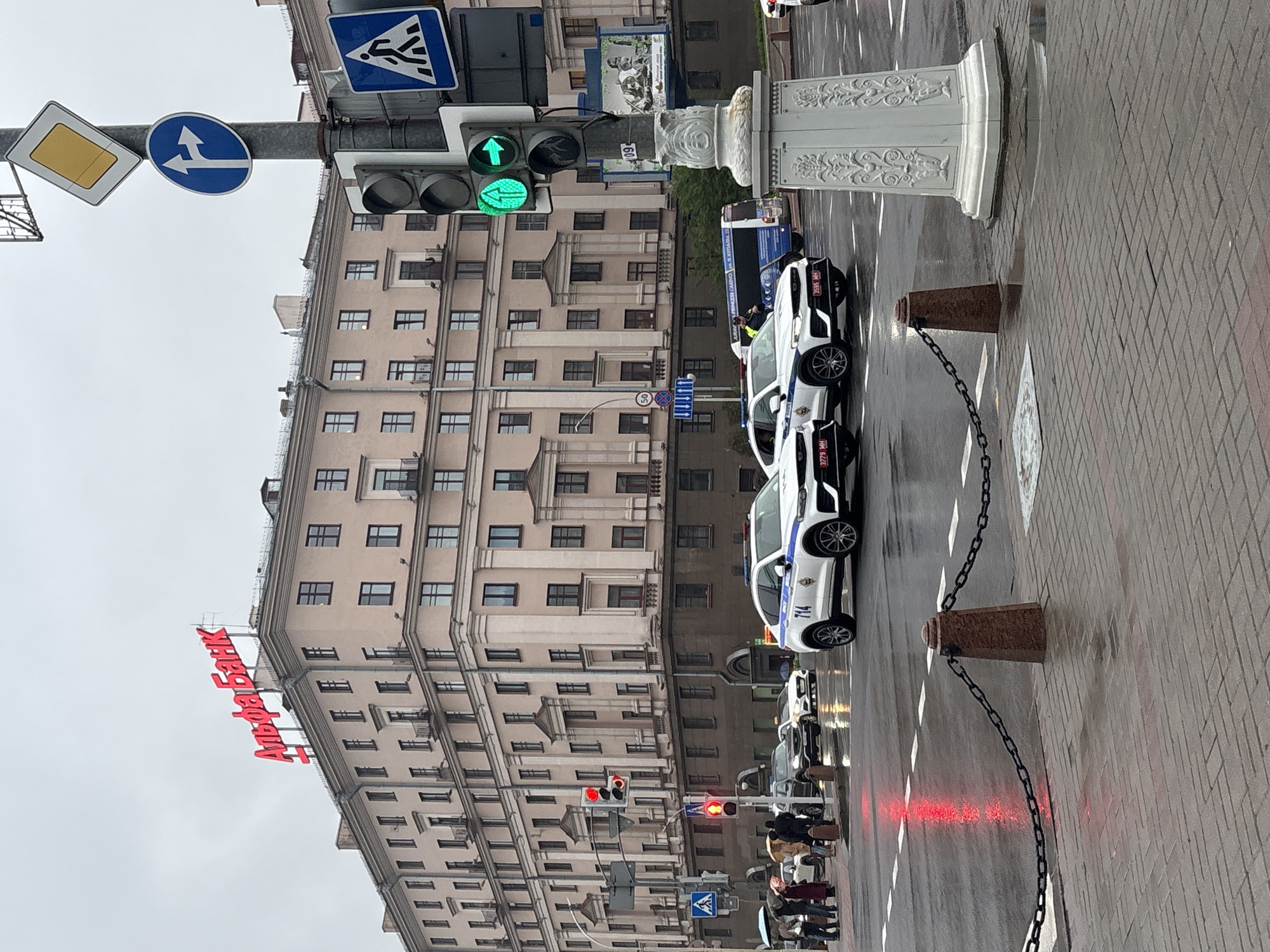 The image depicts a street scene in Minsk, Belarus, under overcast skies. The prominent "Alfa-Bank" sign in red lettering is visible atop a large, multi-story building. Traffic lights display green signals in one direction and red in another. Pedestrians are visible on the sidewalk, some holding umbrellas. A row of white police cars is parked along the curb. Barriers consisting of short pillars linked by chains line the sidewalk. The street is wet, reflecting light.
