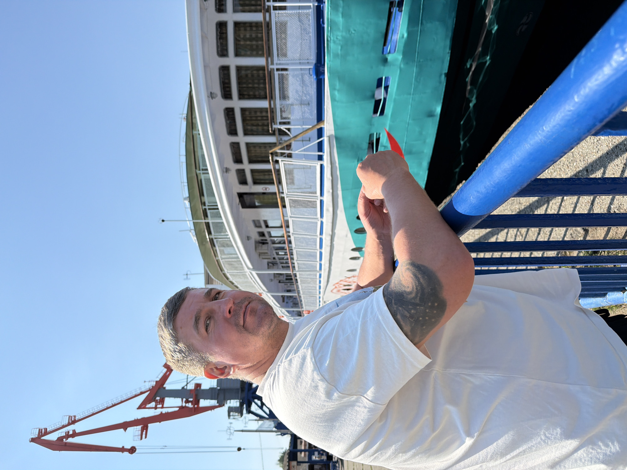 A photograph captures a man in front of a large ship and a crane against a clear sky, possibly in Chaikovsky, Russia. The man, dressed in a white t-shirt, leans against a blue railing with a tattoo visible on his arm. Behind him, a white ship with green and blue accents dominates the background. In the distance, a red and gray crane is also visible. The photo is taken at an angle, giving a dynamic perspective.