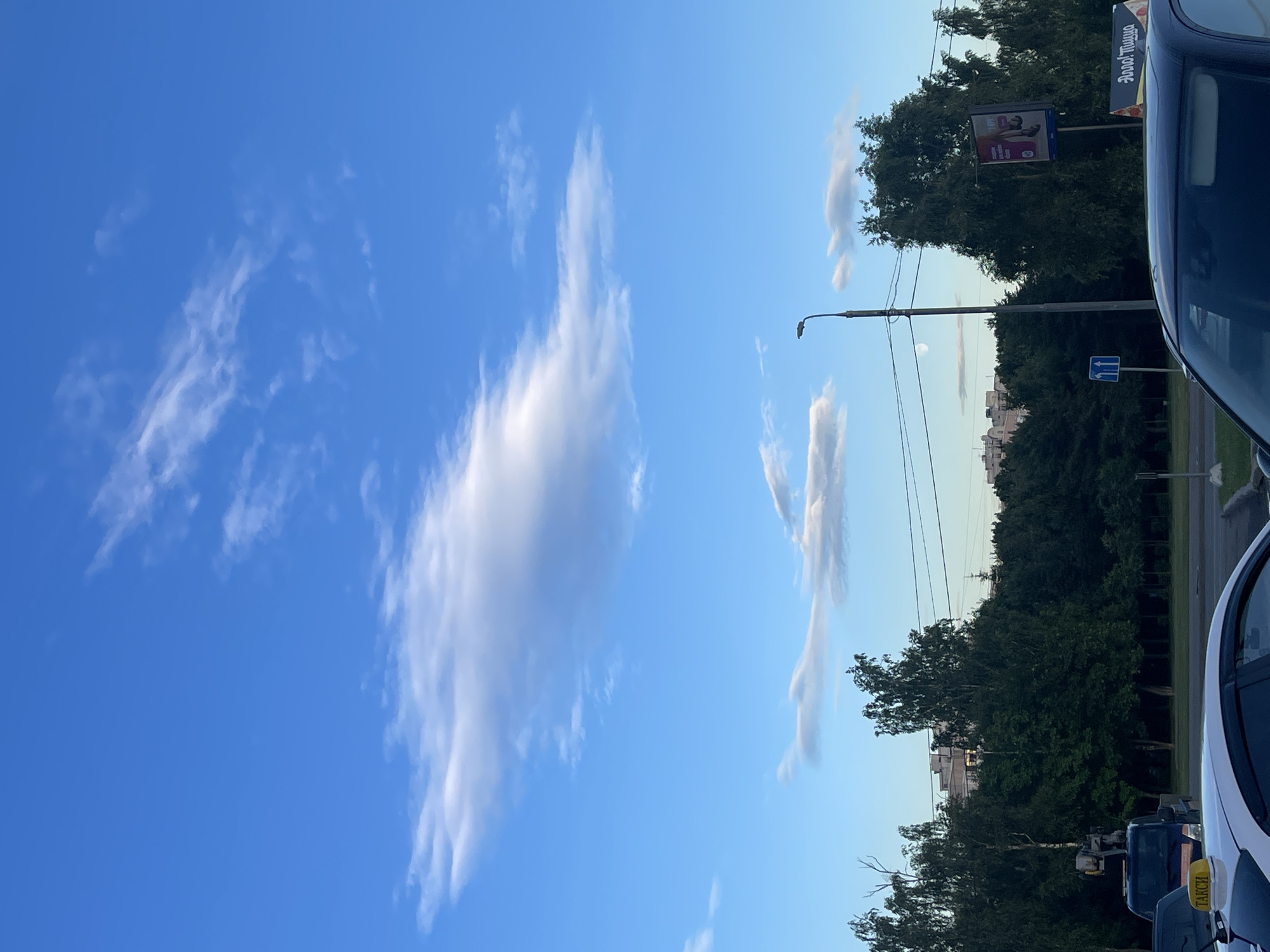 The image captures a scenic view of a bright blue sky with scattered white clouds. In the foreground, the upper portion of a dark blue car is visible on the right side. Behind the car, a dense line of lush green trees forms a backdrop. A tall street lamp stands prominently, with wires stretching across the frame.
Further in the background, several tall buildings can be seen, suggesting an urban or suburban setting. A billboard with an advertisement featuring people is also visible amidst the trees. The lighting suggests it is late afternoon or early evening, with the sun casting a warm glow on the scene. The weather appears clear and pleasant.
On the left side of the image, the sky dominates with its vibrant blue hue and wispy clouds, creating a sense of open space and tranquility. On the right, the car, trees, and buildings ground the scene, providing a sense of place. A sign with blue arrows is visible on a pole. A small yellow sign with the word "ТАКСИ" (Taxi) is affixed to the white car in the foreground. The overall mood is calm and serene, hinting at a peaceful moment in an urban environment.