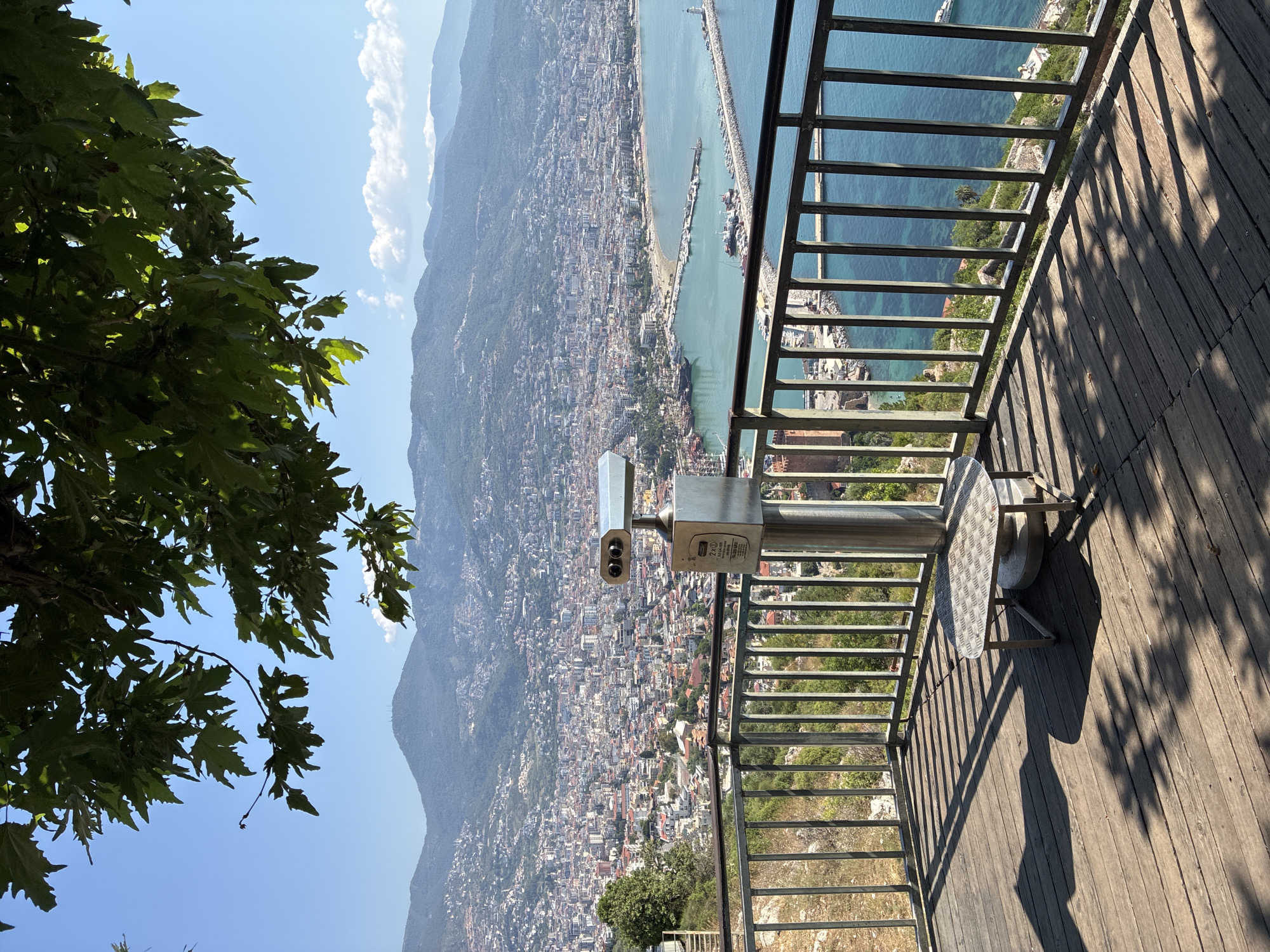 The image captures a scenic viewpoint in Alanya, Turkey, overlooking the expansive city and its coastline. In the foreground, a wooden deck is bordered by a metal railing, under the dappled shadows of tree leaves. A coin-operated binocular viewer stands prominently on the deck, alongside a small, circular metal table. Beyond the railing, the city of Alanya unfolds, a dense tapestry of buildings with red-tiled roofs stretching towards a bay. The bay is dotted with boats and features a long pier extending into the clear blue water. In the distance, rolling hills and mountains form a majestic backdrop under a bright blue sky with scattered white clouds. The overall scene conveys a sense of panoramic beauty and a popular tourist spot offering a commanding view of the city.