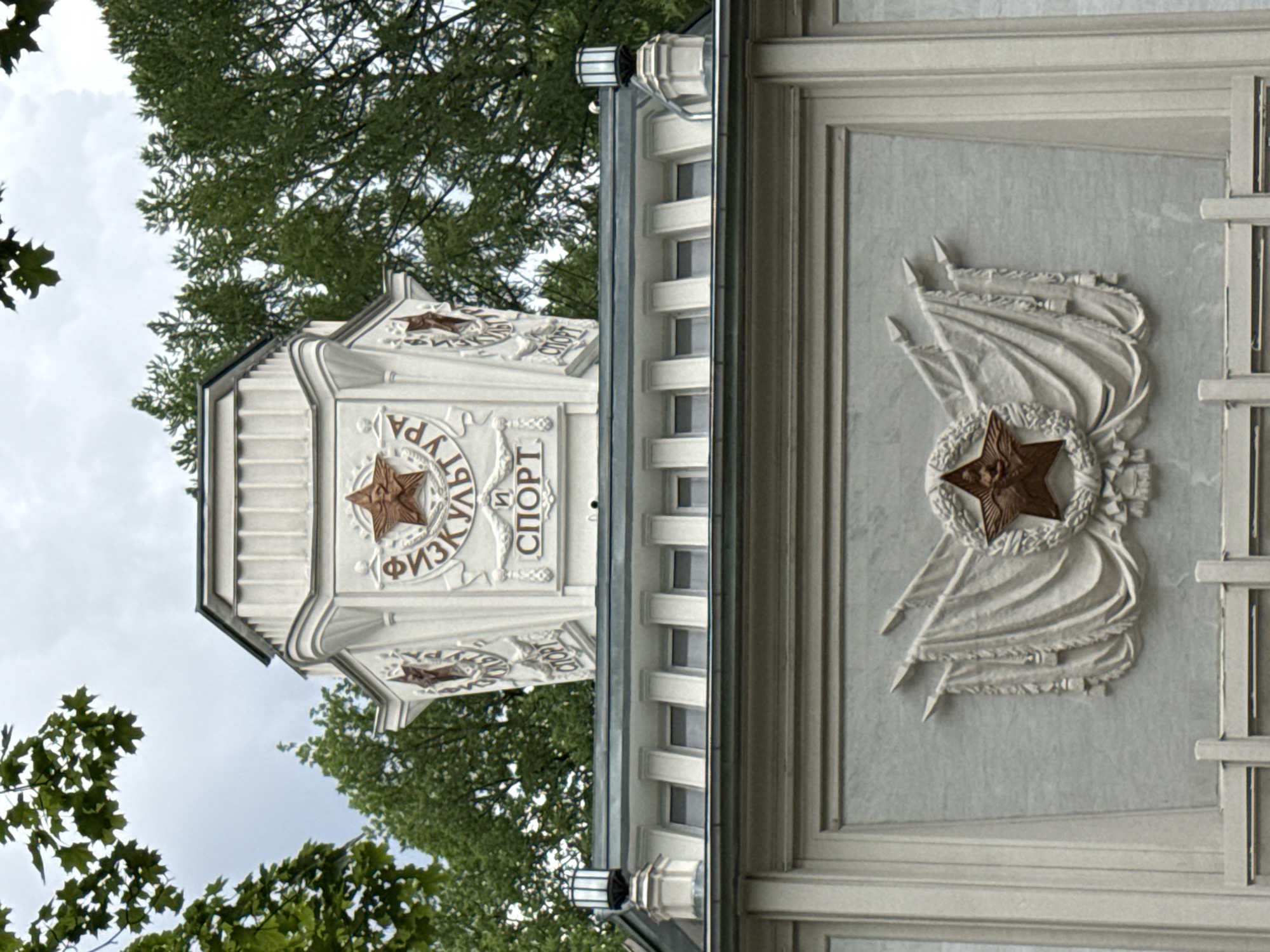 This is a medium-angle, exterior, full-shot photograph of a Soviet-era pavilion at VDNKh (Exhibition of Achievements of National Economy) in Moscow, Russia. The pavilion, painted in a light, off-white color, is adorned with Soviet symbols, including red stars and hammer and sickle emblems. The architecture features ornate reliefs, ribbed columns, and decorative friezes. Two lamps with cylindrical glass shades are visible on the roofline. The sky is overcast, and the tops of green trees frame the upper portion of the structure. The relief on the pavilion's facade includes Cyrillic lettering that reads "ФИЗКУЛЬТУРА И СПОРТ" (Physical Culture and Sport), along with a red star emblem surrounded by a laurel wreath and flags. The lower portion of the image shows another relief with flags and a red star emblem. The overall impression is one of monumental Soviet propaganda architecture.