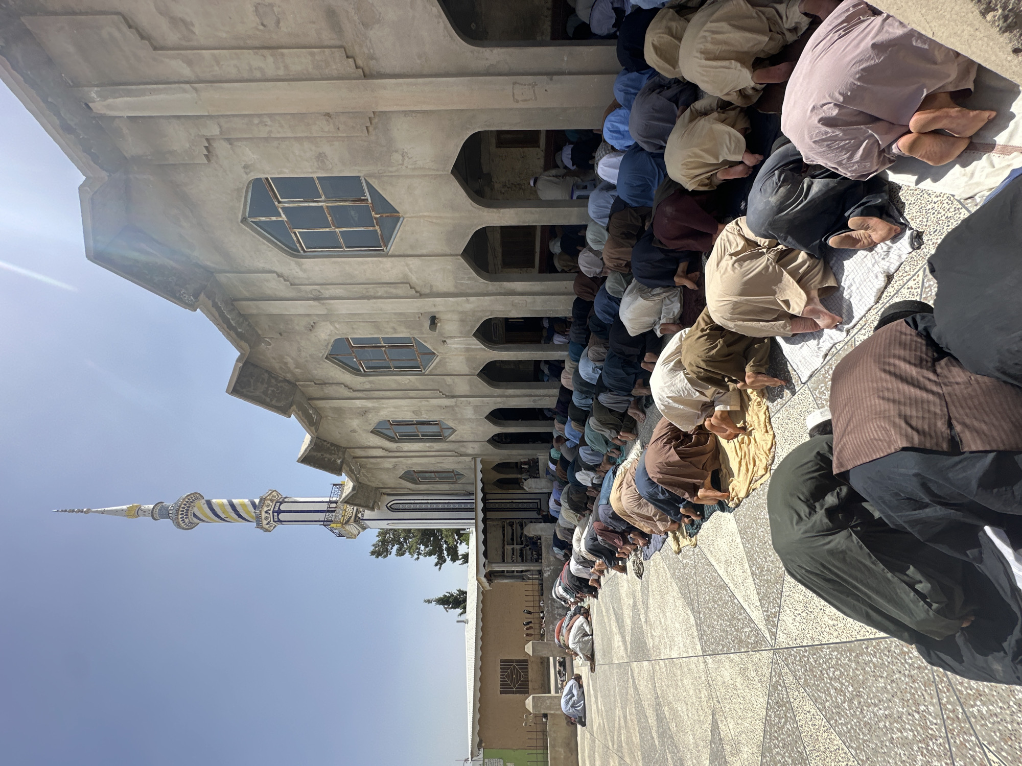 In an outdoor courtyard of a mosque, a large group of men are engaged in communal prayer. They are arranged in neat rows, facing the mosque, and are in various stages of prayer, primarily in a prostrated position. The men are dressed in traditional Muslim attire, with many wearing long tunics and head coverings in a variety of earth tones, whites, and blues. The courtyard pavement is a geometric pattern of light-colored tiles. To the right, the mosque building is visible, constructed of concrete with arched doorways and square-paned windows. A prominent, colorful minaret with blue, yellow, and white spiral decorations rises above the mosque on the left side of the frame, against a clear blue sky. The sun is bright, casting shadows on the ground, and the overall scene suggests a religious gathering under a warm, sunny sky.