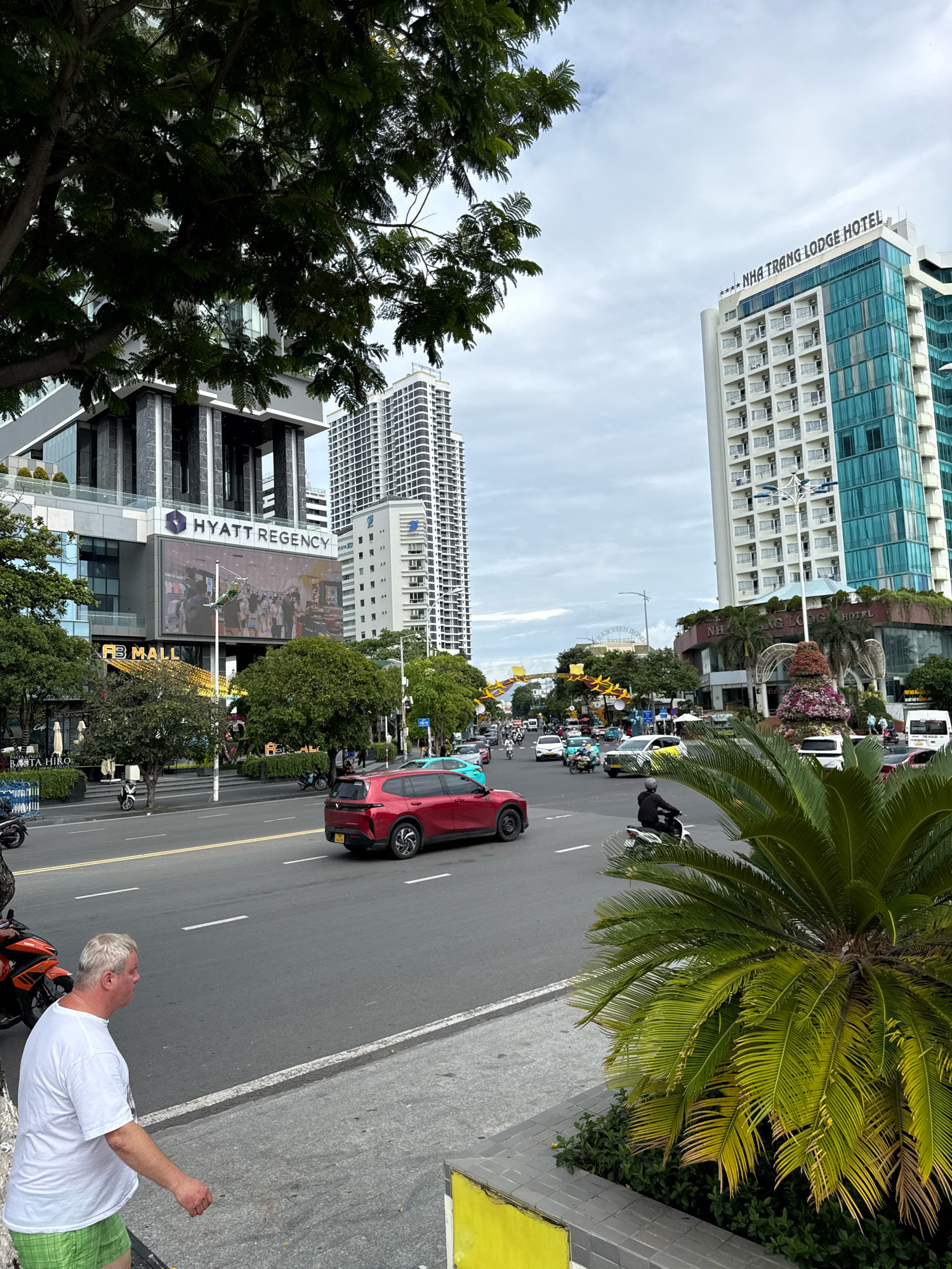 The image depicts a street scene in Nha Trang, Vietnam, with multiple buildings lining the road. In the foreground, a white man wearing a white t-shirt and green shorts walks on a sidewalk. To his left, an orange motorcycle is parked. The street is busy with traffic, including a red SUV, several other cars, and motorcycles. Buildings visible include the Hyatt Regency and the Nha Trang Lodge Hotel, with one prominent building featuring a large digital billboard. Lush green trees and palm fronds are interspersed throughout the urban landscape. The sky is overcast.