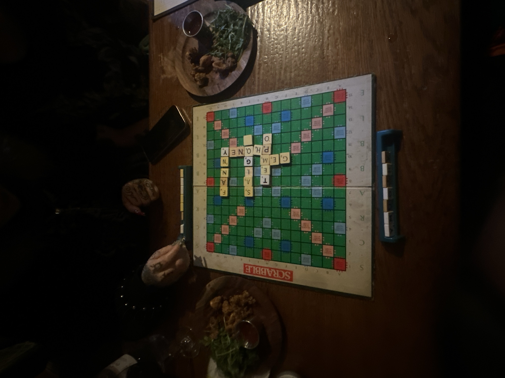 A photograph shows a Scrabble board game set up on a wooden table, positioned alongside dishes containing food items. The board features the word "PHONEY" spelled out horizontally. Near the board, there are scattered food dishes, including what appears to be fried food and leafy greens. The table is made of a dark wood, providing a warm backdrop to the scene. The environment suggests an informal setting, such as a game night at home or a casual gathering at a pub or restaurant, but there are no identifying details of a specific location in London, UK. Visible are partially obscured persons; their clothing and visible skin features hands with tattoos, and accessories such as studded choker necklaces.