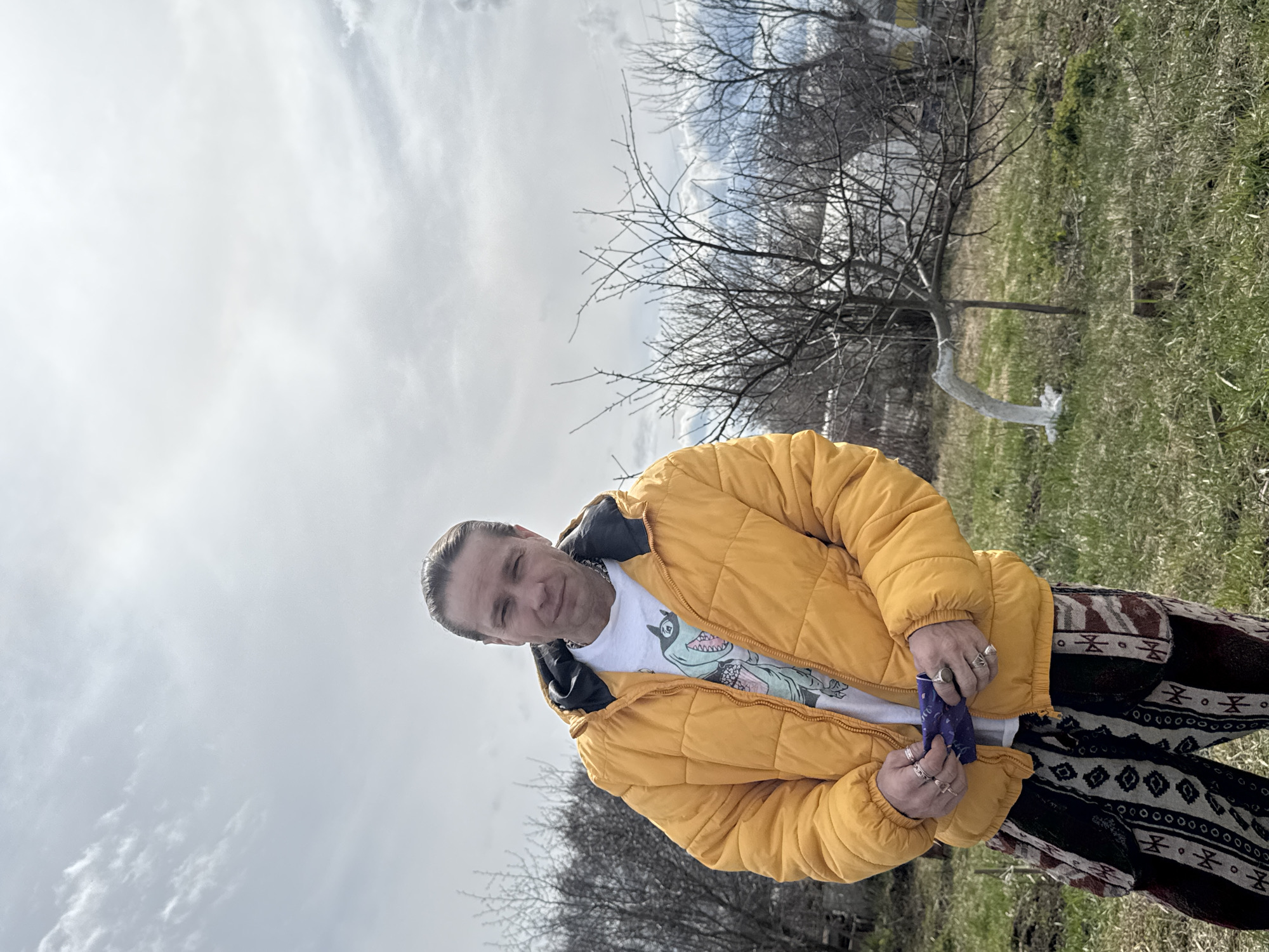 An outdoor portrait captured in Chaikovsky, Russia, features a man standing with a cloudy sky overhead. The man, with light-colored hair pulled back, looks directly at the viewer with a neutral to slight smiling expression. He is wearing a bright yellow, puffy winter jacket with the zipper slightly open, revealing a white t-shirt underneath. The t-shirt has a colorful graphic depicting a dinosaur, and the partially visible text "T-Rex" can be seen on it. He also wears dark pants with a distinctive white and brown geometric patterned design. Rings are visible on several fingers of both hands, and he appears to be holding a small, dark blue, crinkled item in his right hand.
The background consists of a natural setting with bare trees, indicating late winter or early spring. The ground is covered with green grass. One tree trunk in the mid-ground appears to be painted white near its base. The sky is overcast with visible white and gray clouds, suggesting a bright but sunless day. The overall scene conveys a casual outdoor moment.