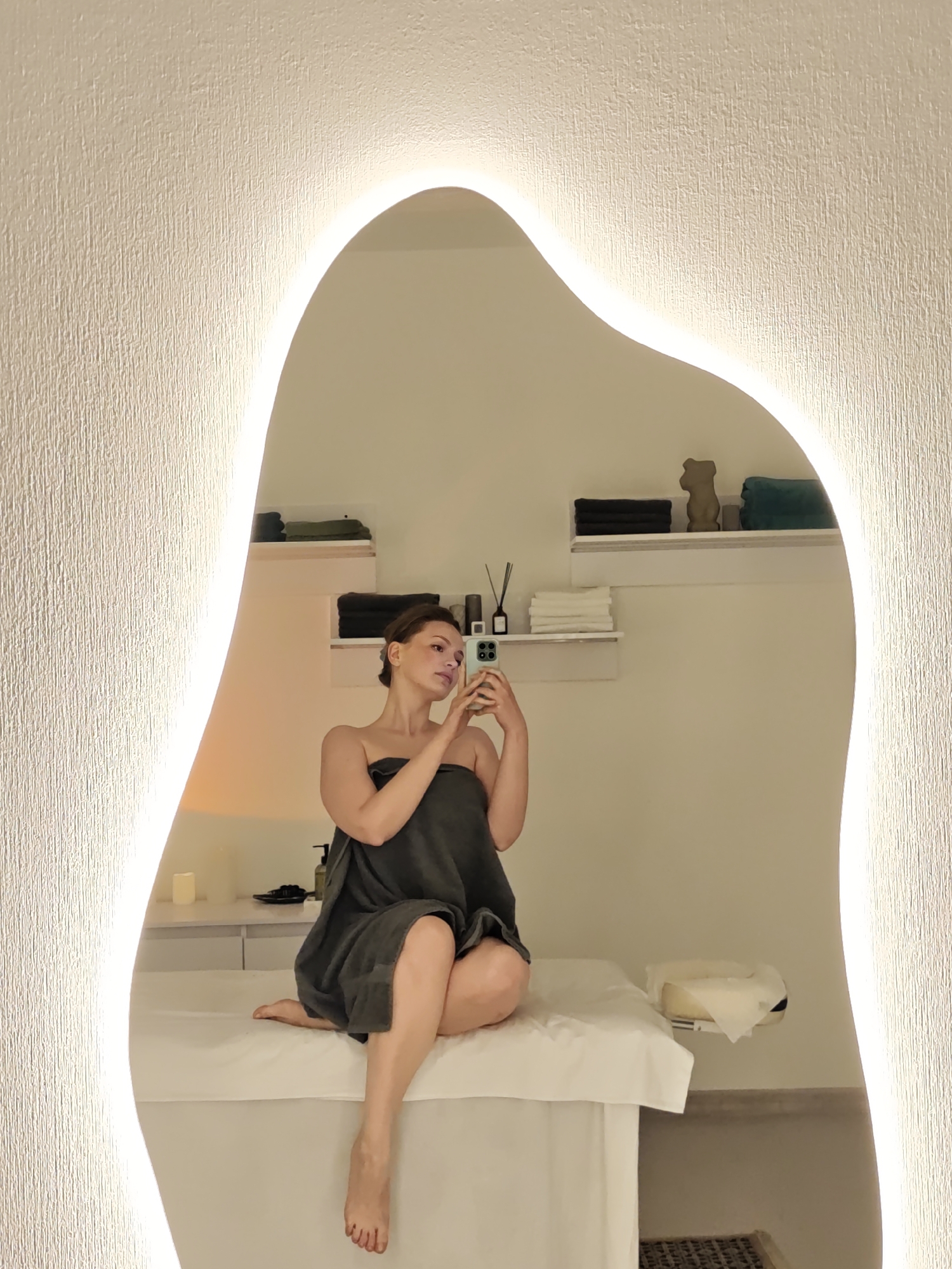 A woman is sitting on a massage table in a spa. She is wrapped in a dark gray towel and holding a smartphone up to take a selfie in a large, irregularly shaped mirror with a lighted border. The spa room is decorated in neutral tones with white walls and furniture. There are shelves holding folded towels, a teddy bear figure, and decorative reed diffusers. A candle and lotion bottles are visible on a countertop. The location of the photo, if taken in Vinnytsia, Ukraine, cannot be determined definitively from the image without additional contextual information.
