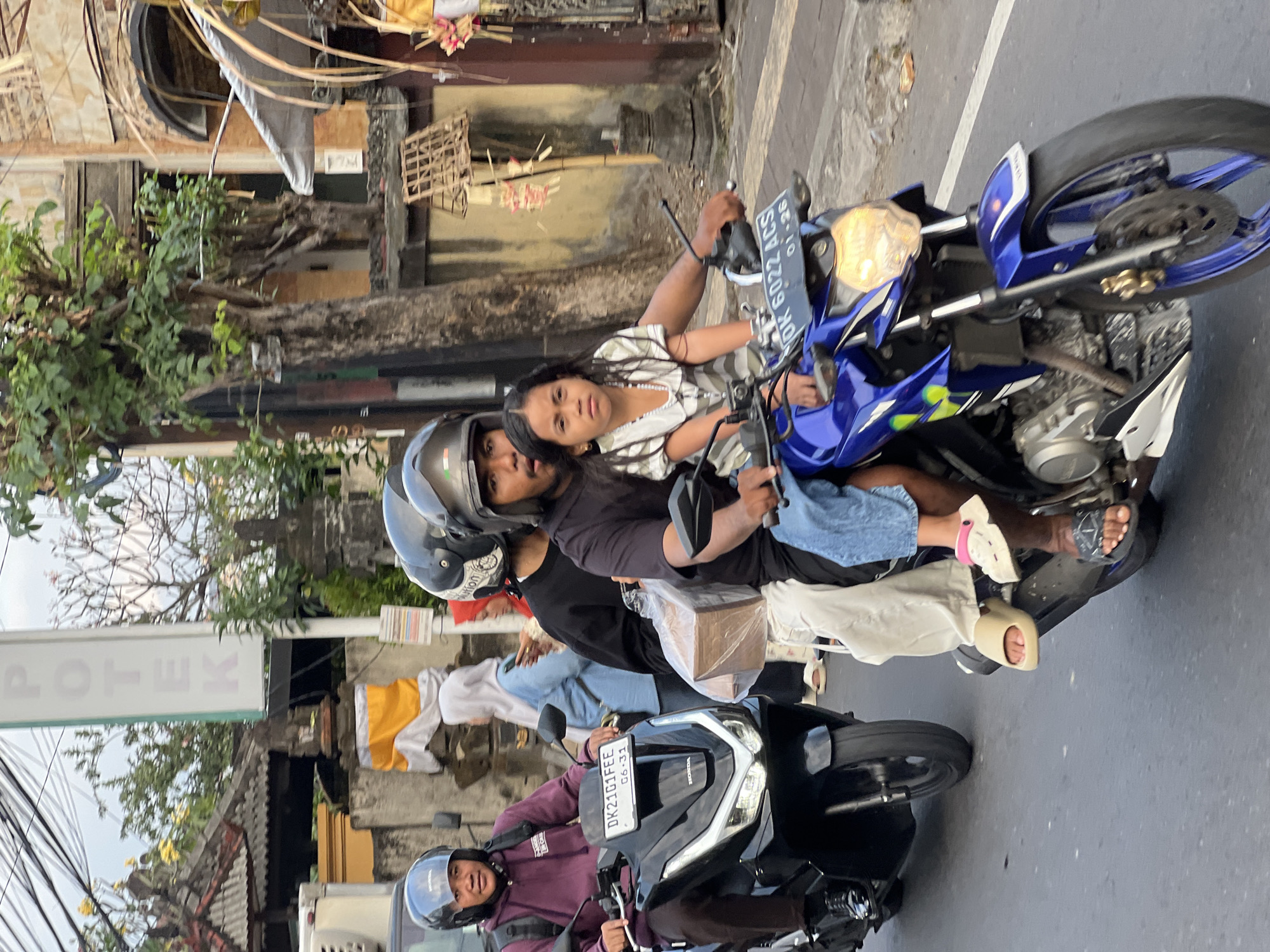 On a street in Denpasar, Indonesia, three people, a woman and two children, ride on a blue motorcycle. The woman, wearing a black t-shirt, drives the motorcycle. A young girl, wearing a white and green striped dress and a necklace, sits in front of the woman. Another person, wearing a black t-shirt and a white headscarf, sits behind the girl, holding a brown box wrapped in plastic. The motorcycle is traveling down a paved street. In the background, there are buildings and trees. The sky is overcast. A white sign with the word "APOTEK" is visible on the left side of the image. Another motorcycle is visible on the left side of the image.