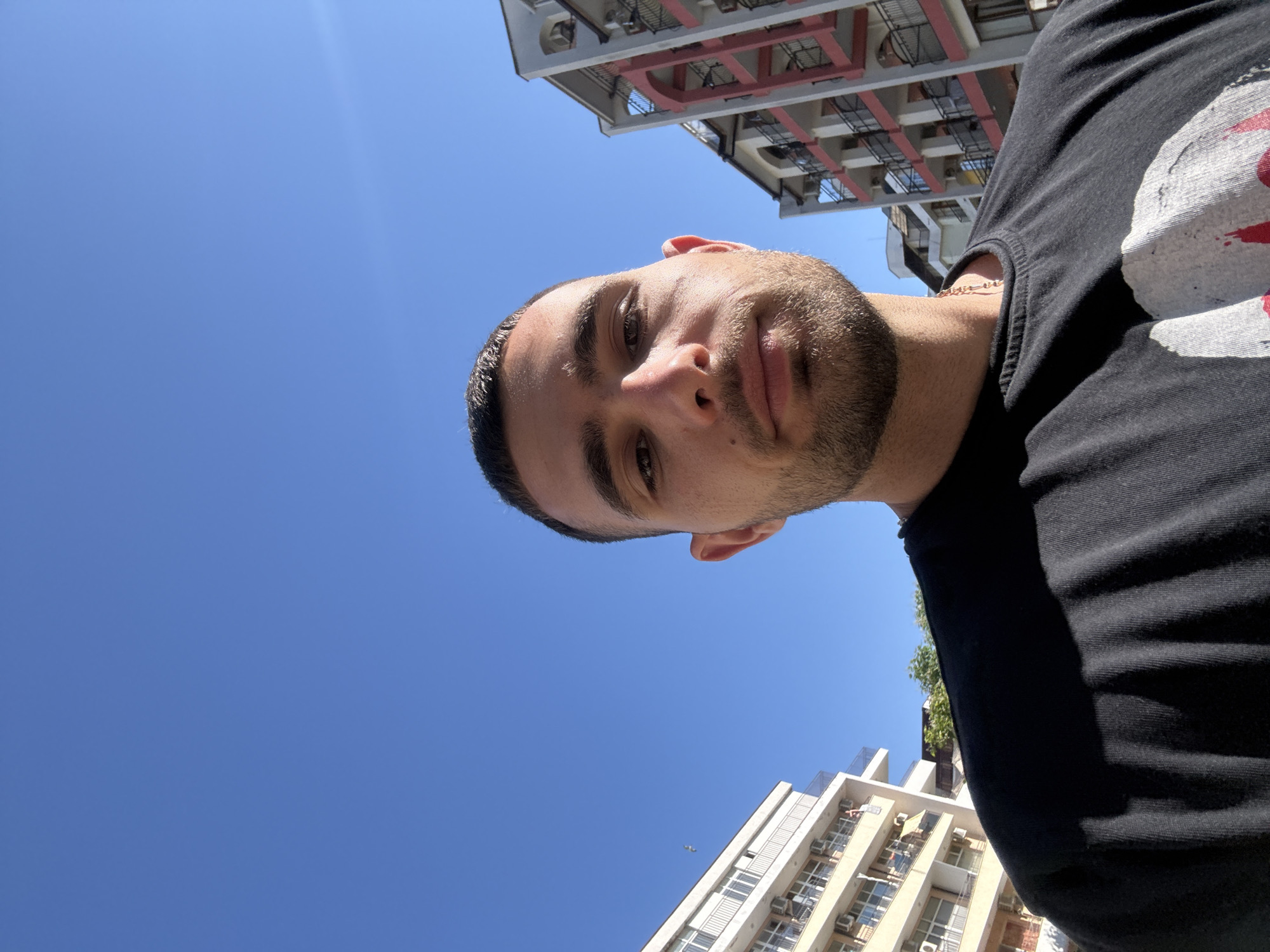 A young man with short dark hair and stubble is pictured from the chest up, facing the camera. He is wearing a black t-shirt. The background is a clear blue sky with two apartment buildings on either side of the frame. The building on the left is light-colored with white balconies, and the building on the right is pink and white with red balconies. The scene appears to be in Nesebar, Bulgaria, a coastal city known for its historic old town and modern beach resorts.